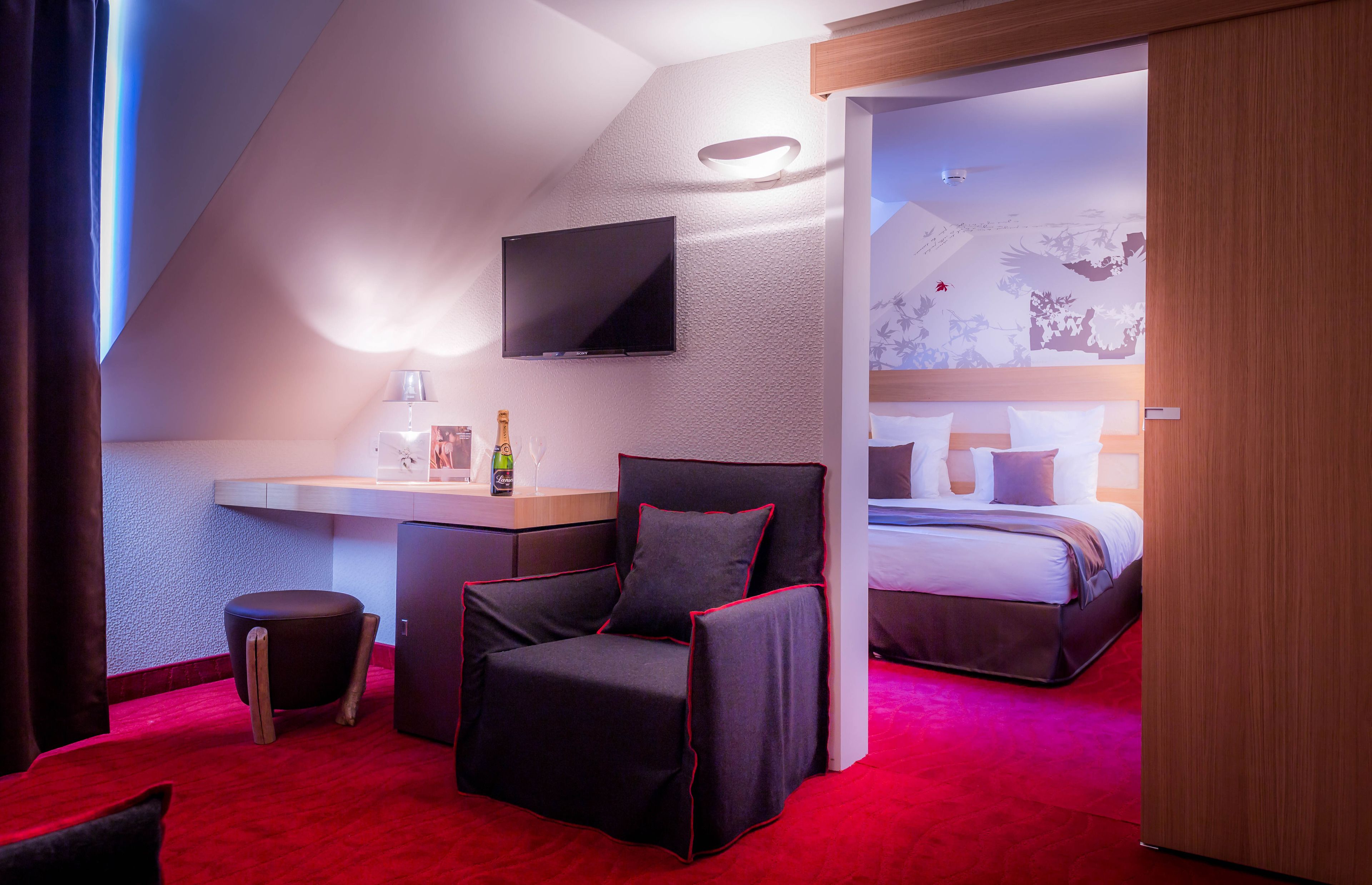 Une chambre d’hôtel moderne équipée d’une télévision à écran plat, d’un bureau avec une lampe, d’un fauteuil confortable, d’un lit avec une tête de lit et d’une moquette rouge.