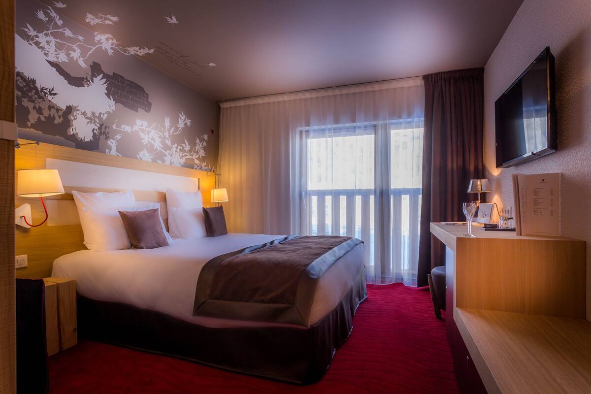 A hotel room with a large bed, a desk with a flat-screen TV, and a window with curtains.
