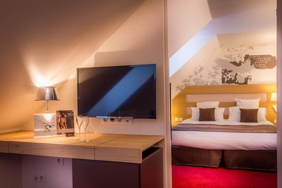 A hotel room with a flat-screen TV, a desk with a lamp, and a bed with a red carpet.