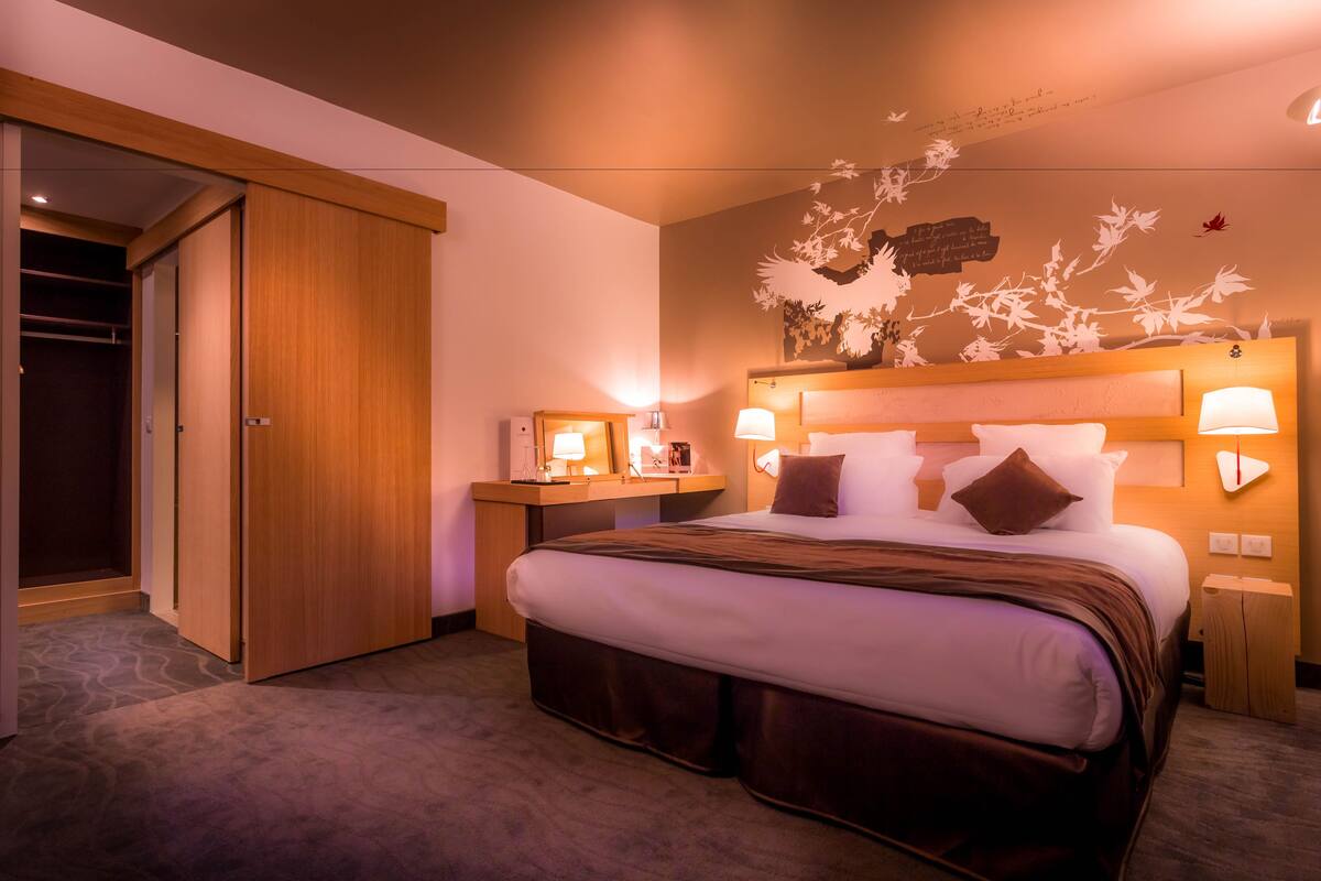 A hotel room with a large bed, wooden headboard, bedside lamps, a nightstand, and a wardrobe.