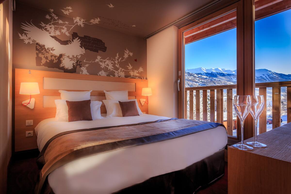 A hotel room with a large bed, wooden headboard, and a view of snow-capped mountains through a balcony.