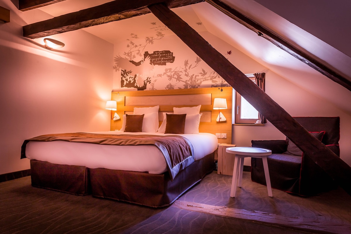 A bedroom with a large bed, bedside lamps, a small table, and a sloped ceiling with exposed beams.