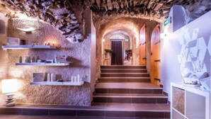Un pasillo de paredes de piedra con una escalera que conduce a una puerta al final. Se ven estantes con objetos en la pared izquierda.