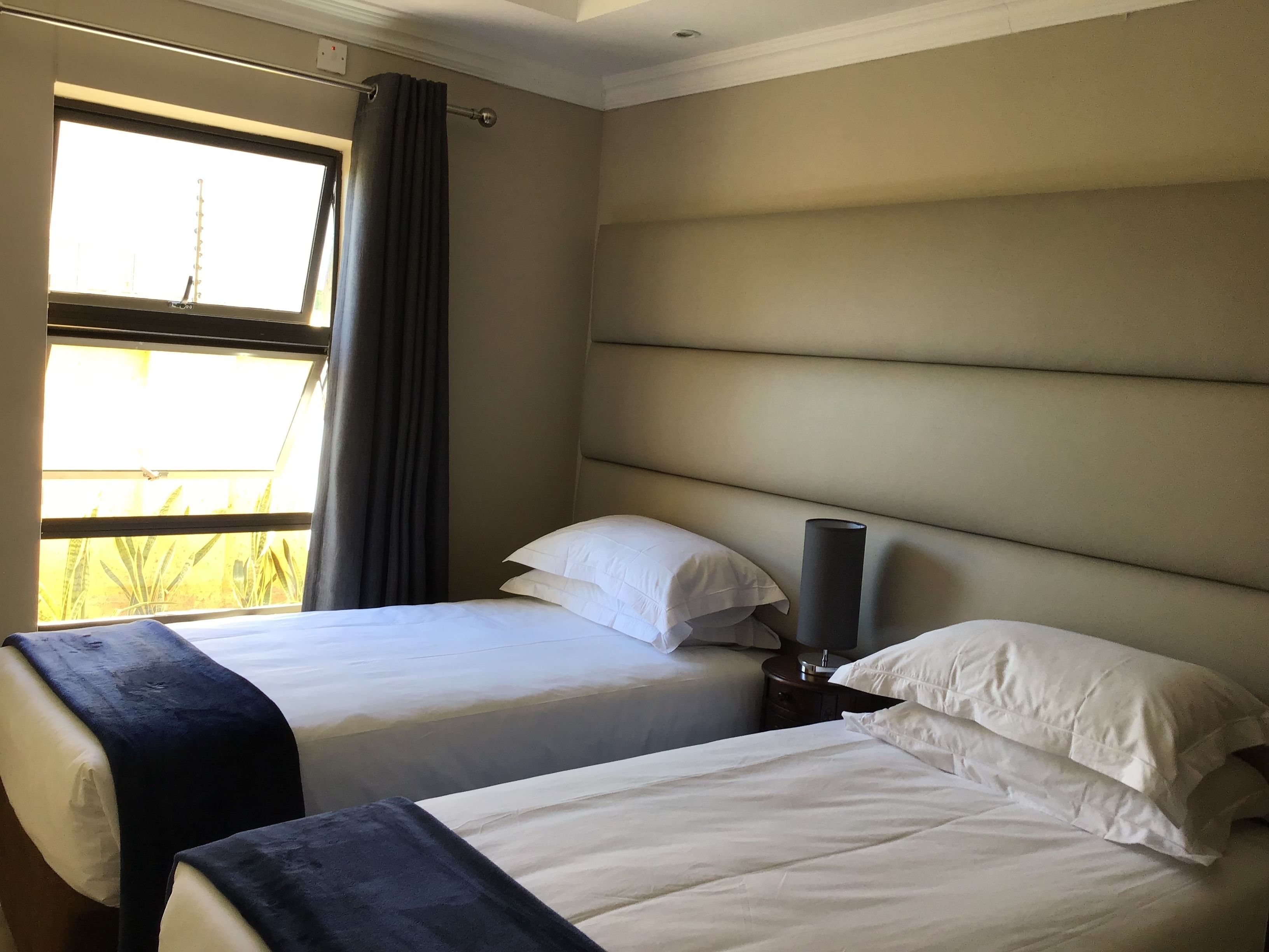 A hotel room with two beds, a window with curtains, a bedside lamp, and a view of greenery outside.