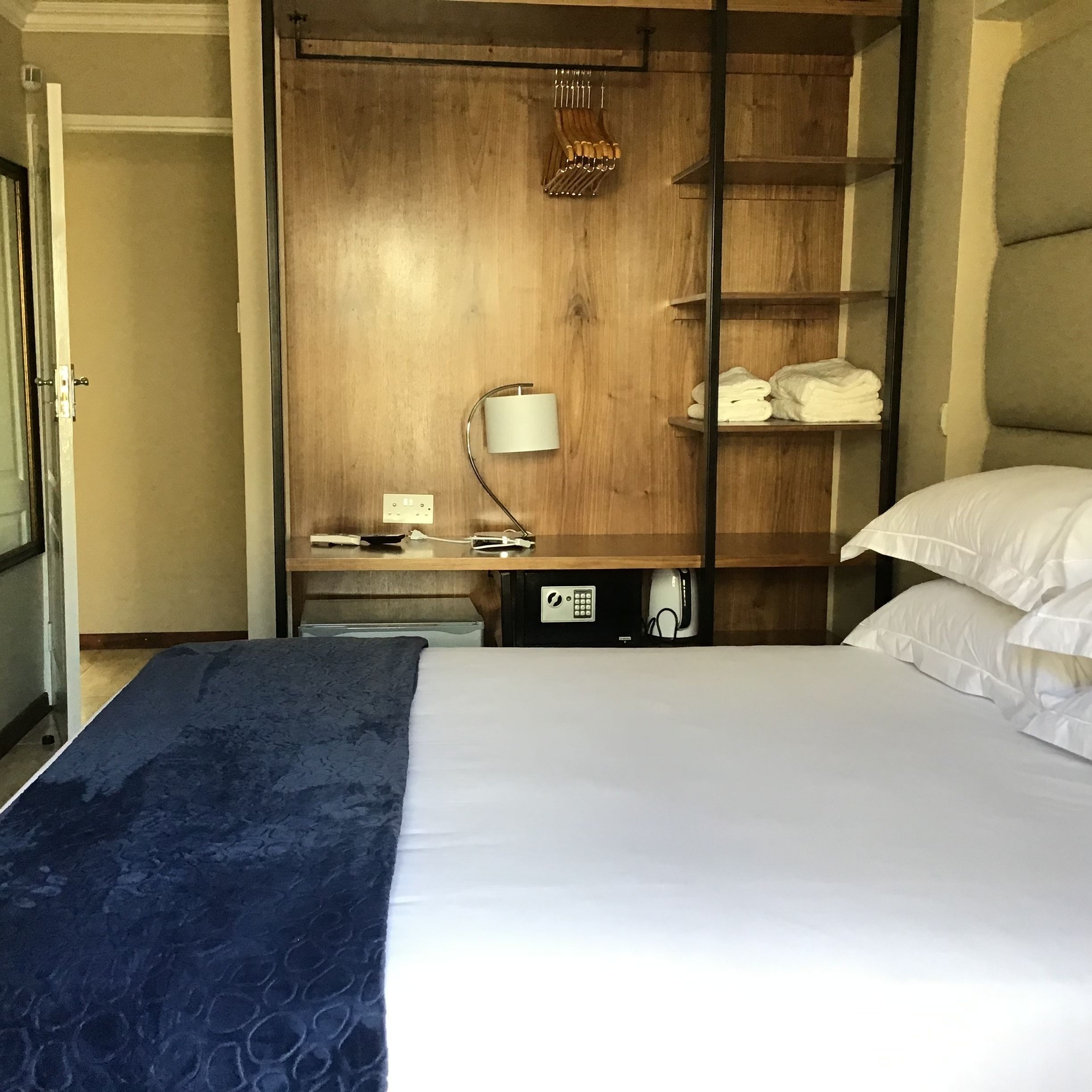 A hotel room with a bed, a desk with a lamp, a wardrobe, and a shelf with towels.
