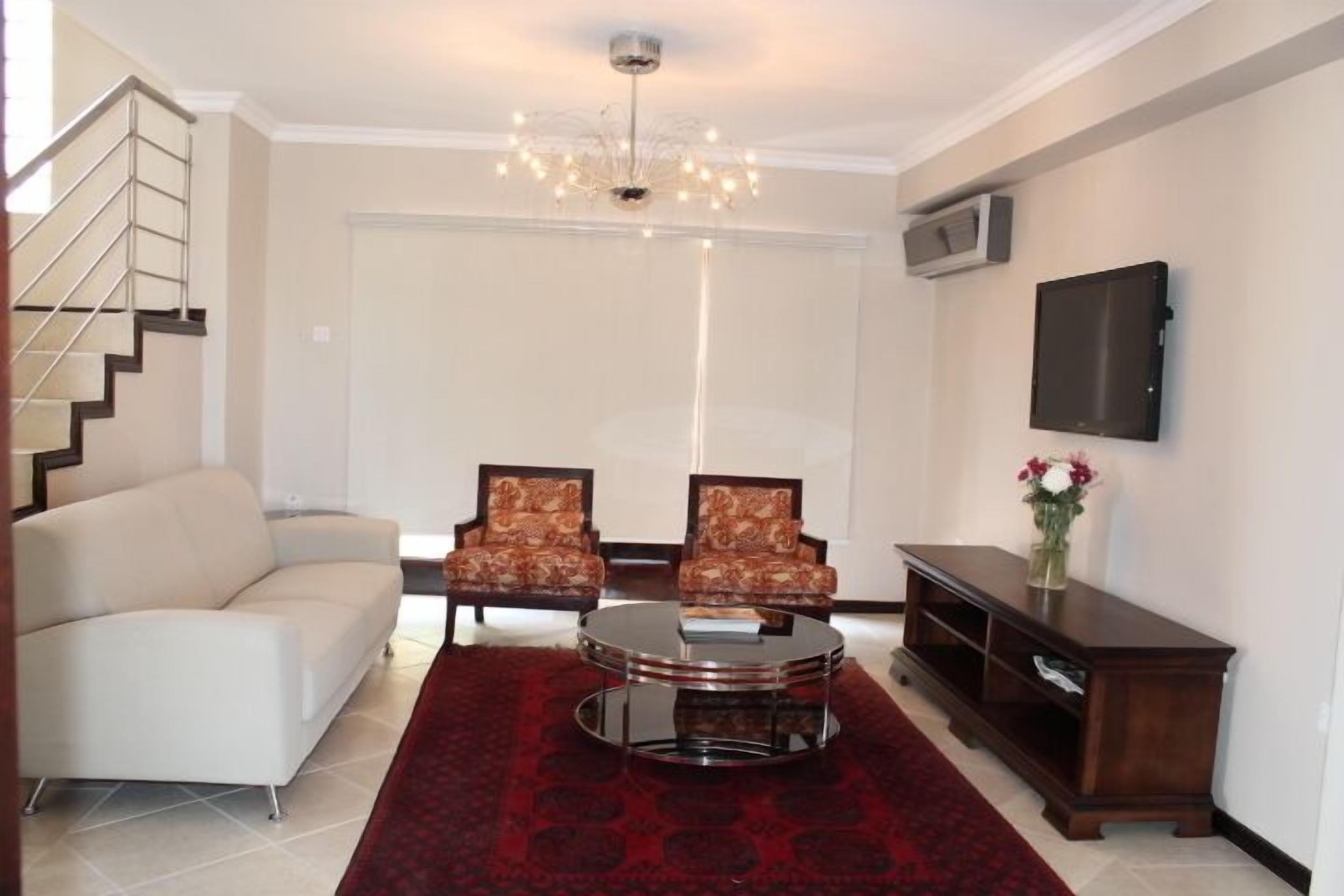 A living room with a sofa, two patterned armchairs, a round coffee table, a red rug, a TV on the wall, and a staircase.