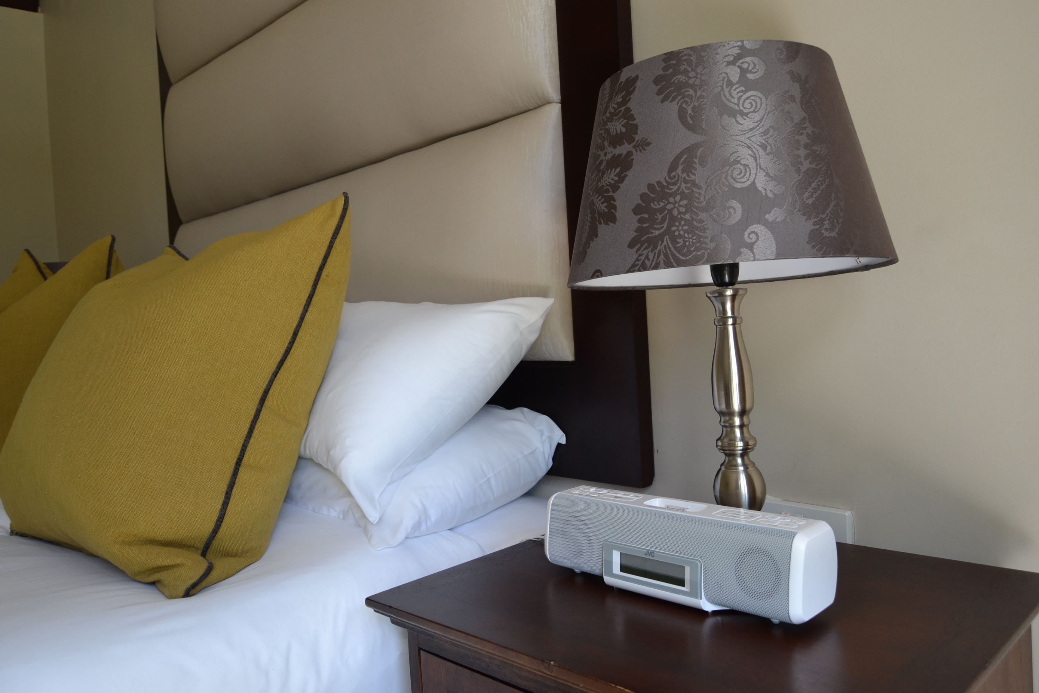 A neatly made bed with white and mustard pillows, a bedside table with a lamp, and a white electronic device on the table.