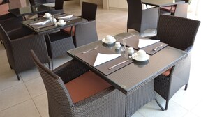 A dining area with wicker furniture, glass tables, and neatly arranged table settings. - The Capital Guesthouse (Gaborone)