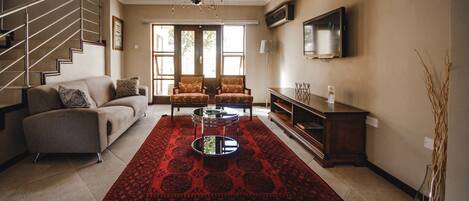 A living room with a staircase, a sofa, two armchairs, a coffee table, and a TV on the wall.