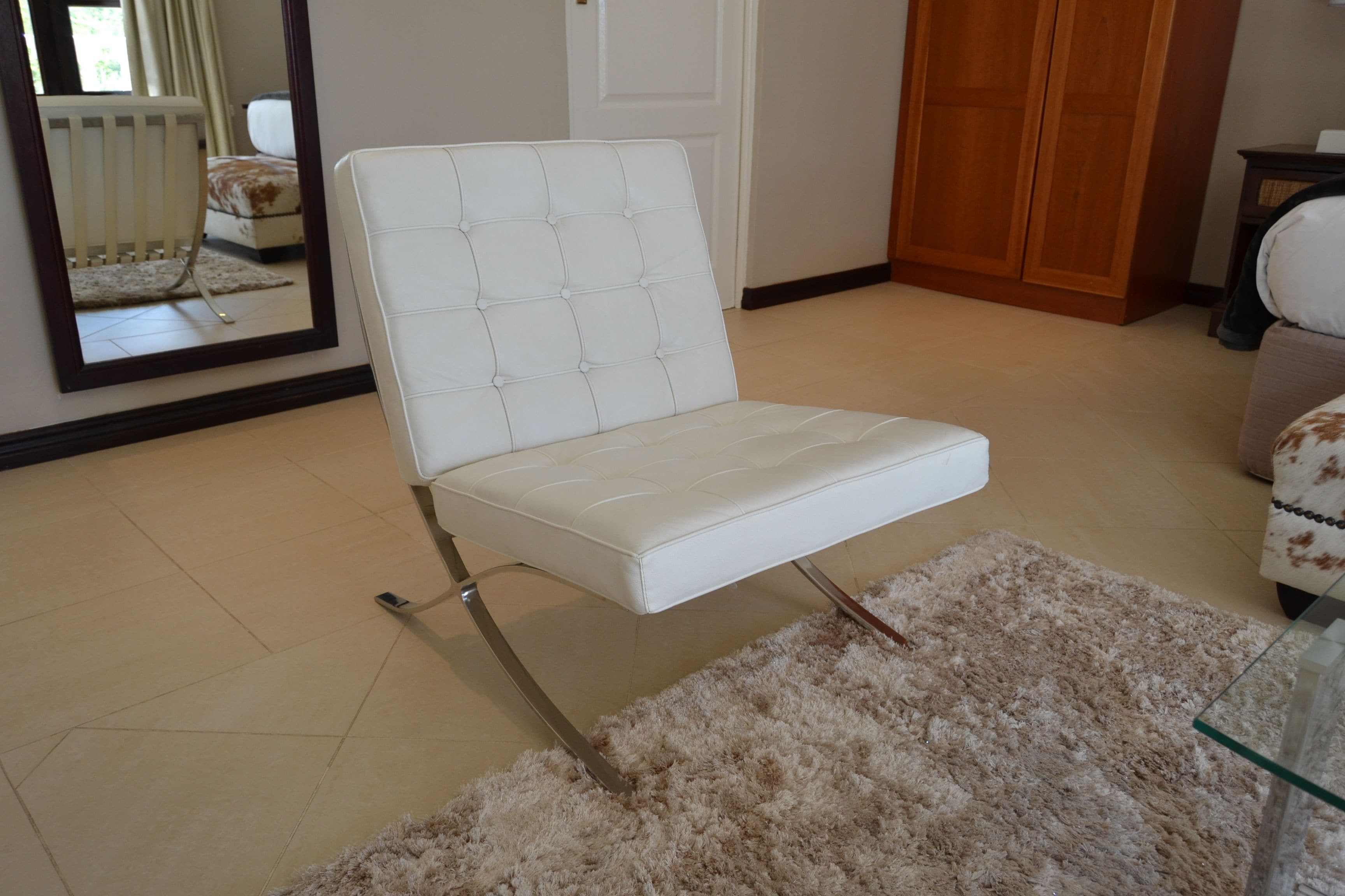 A white cushioned chair with a high backrest and metal legs, positioned on a beige carpet in a room with a wooden wardrobe and a mirror reflecting a crib and a bed.