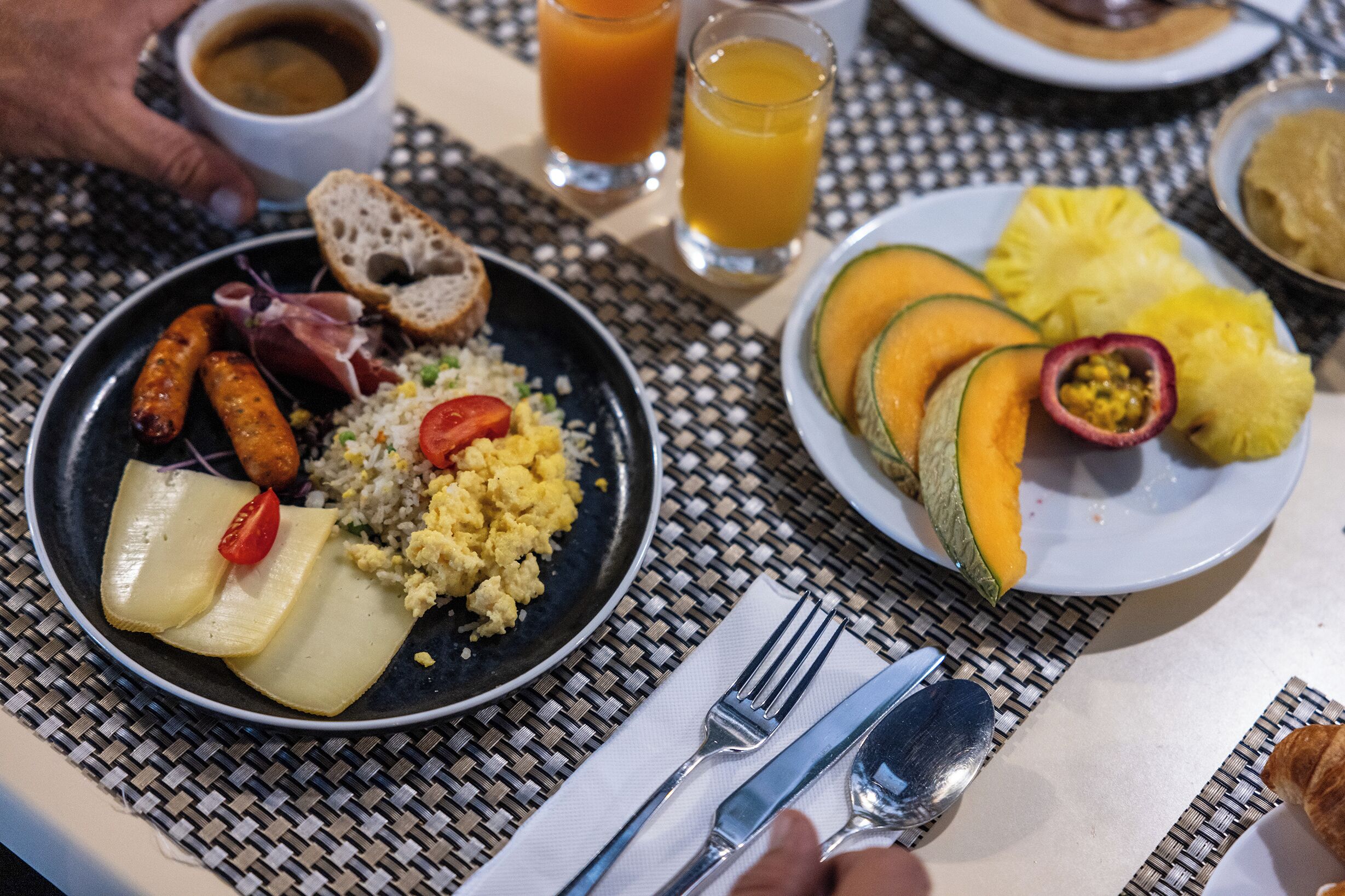 A breakfast setting with plates of food, a cup of soup, and glasses of orange juice on a woven tablecloth.