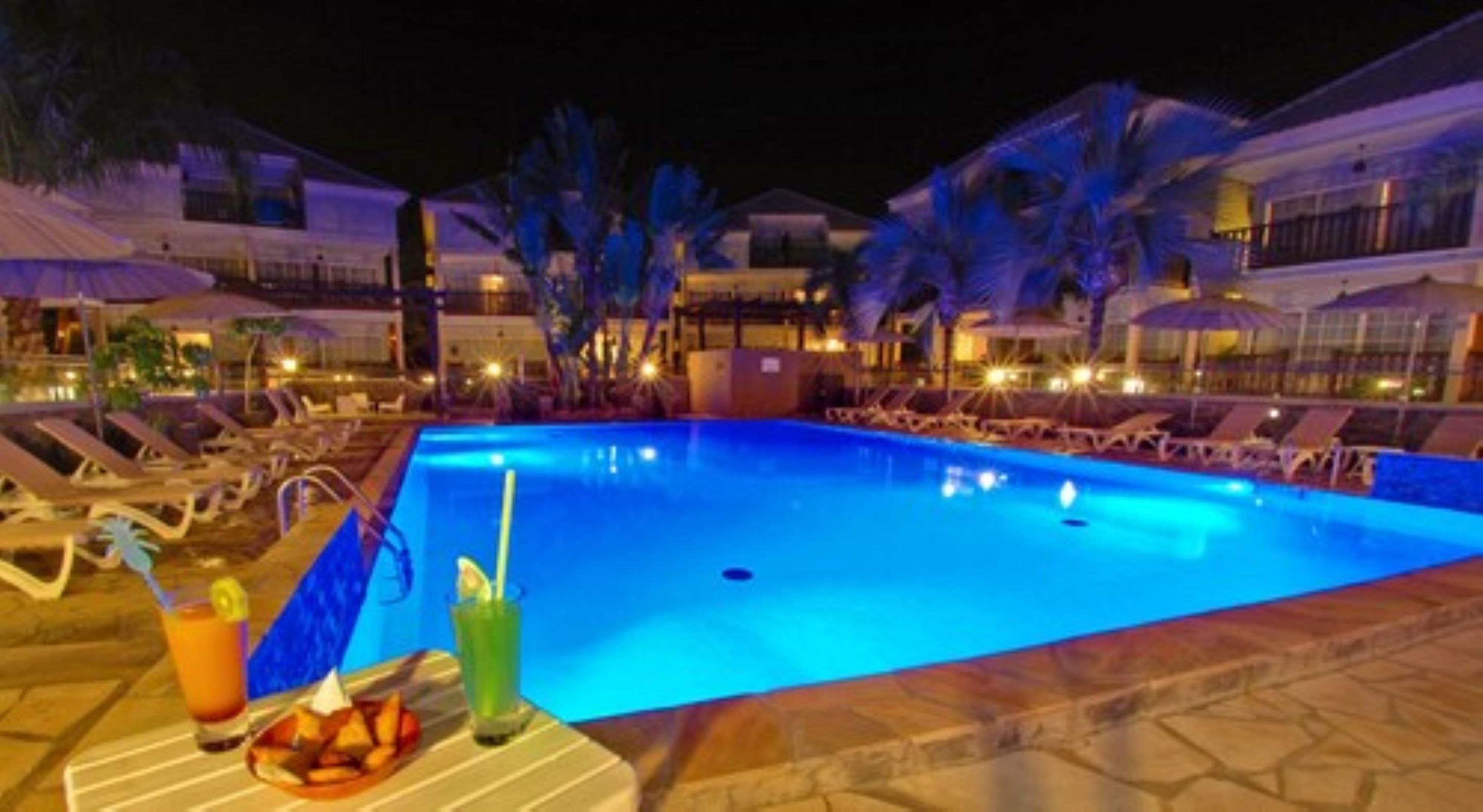 A nighttime swimming pool with lounge chairs and umbrellas, surrounded by buildings with illuminated windows.