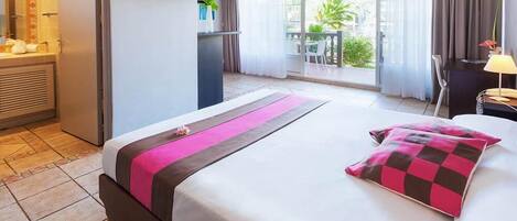 A hotel room with a bed, a TV mounted on the wall, a balcony with a table and chairs, and a view of greenery outside.
