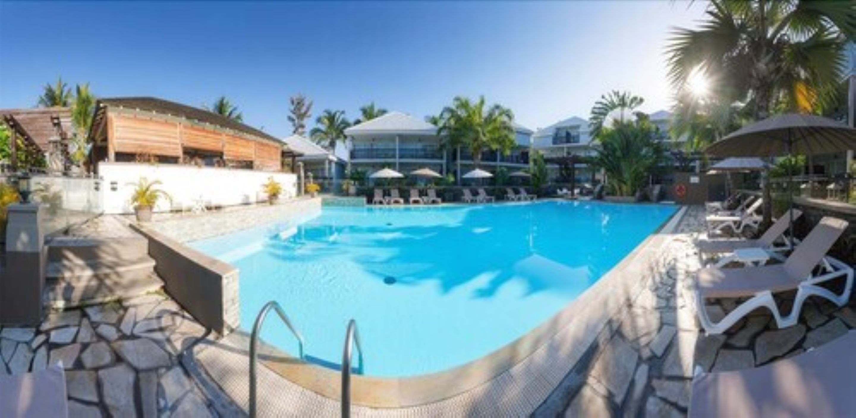 A swimming pool with lounge chairs and umbrellas, surrounded by palm trees and modern buildings.