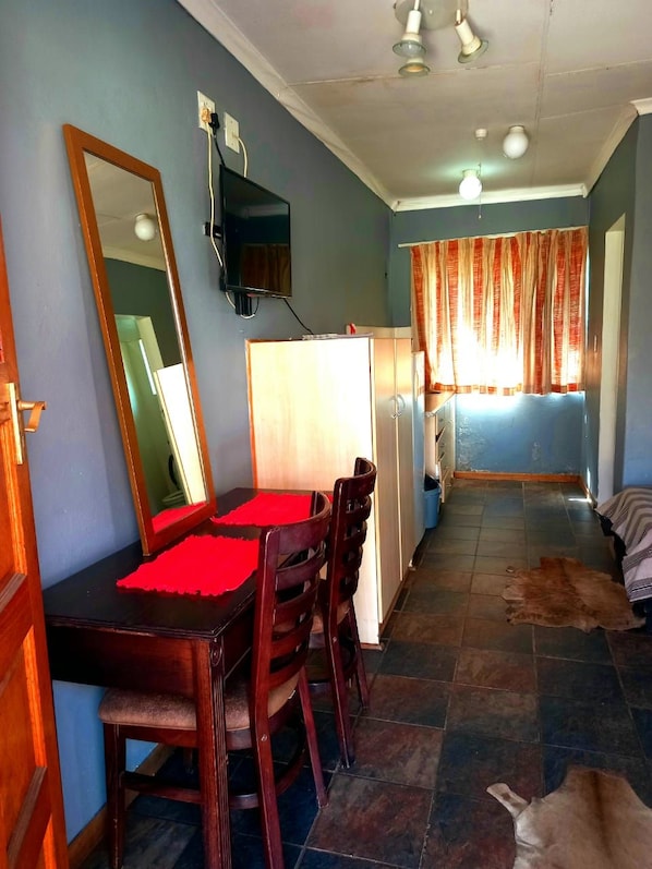 Quarto com mesa de jantar, cadeiras, televisão na parede, janela com cortinas e tapete no chão.