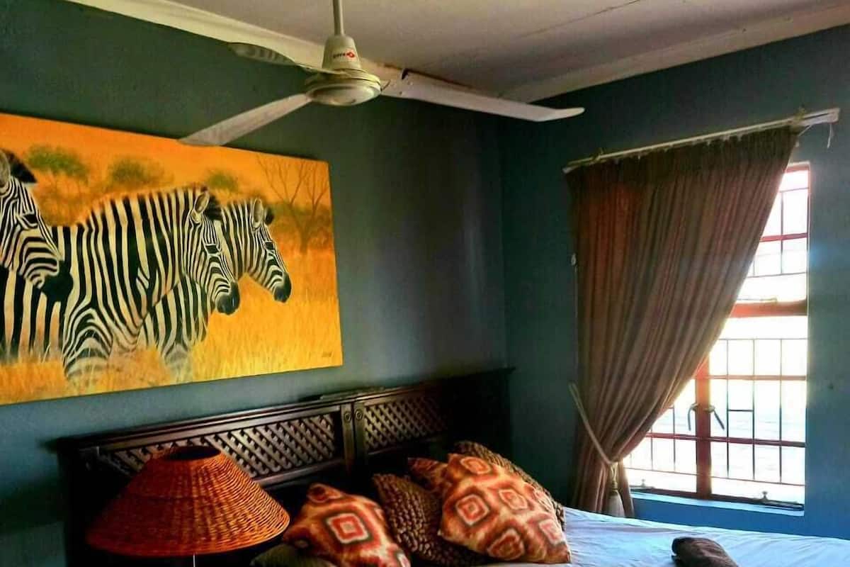Une chambre à coucher avec un lit, une table de chevet, une lampe, un ventilateur de plafond, un tableau représentant une zèbre et une fenêtre avec des rideaux.