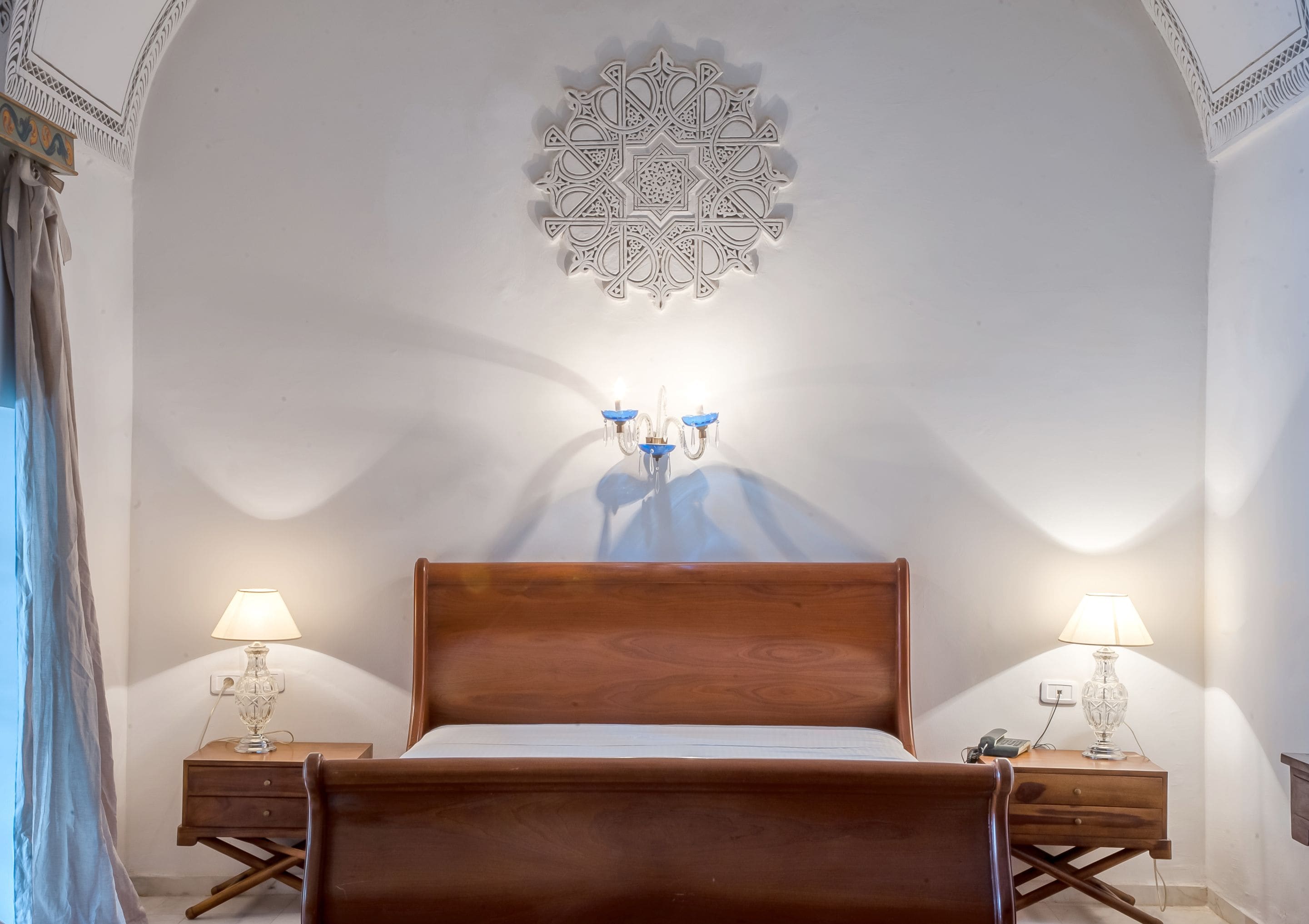 A bedroom with a wooden bed, two bedside tables with lamps, a decorative ceiling medallion, and a wall-mounted light fixture.