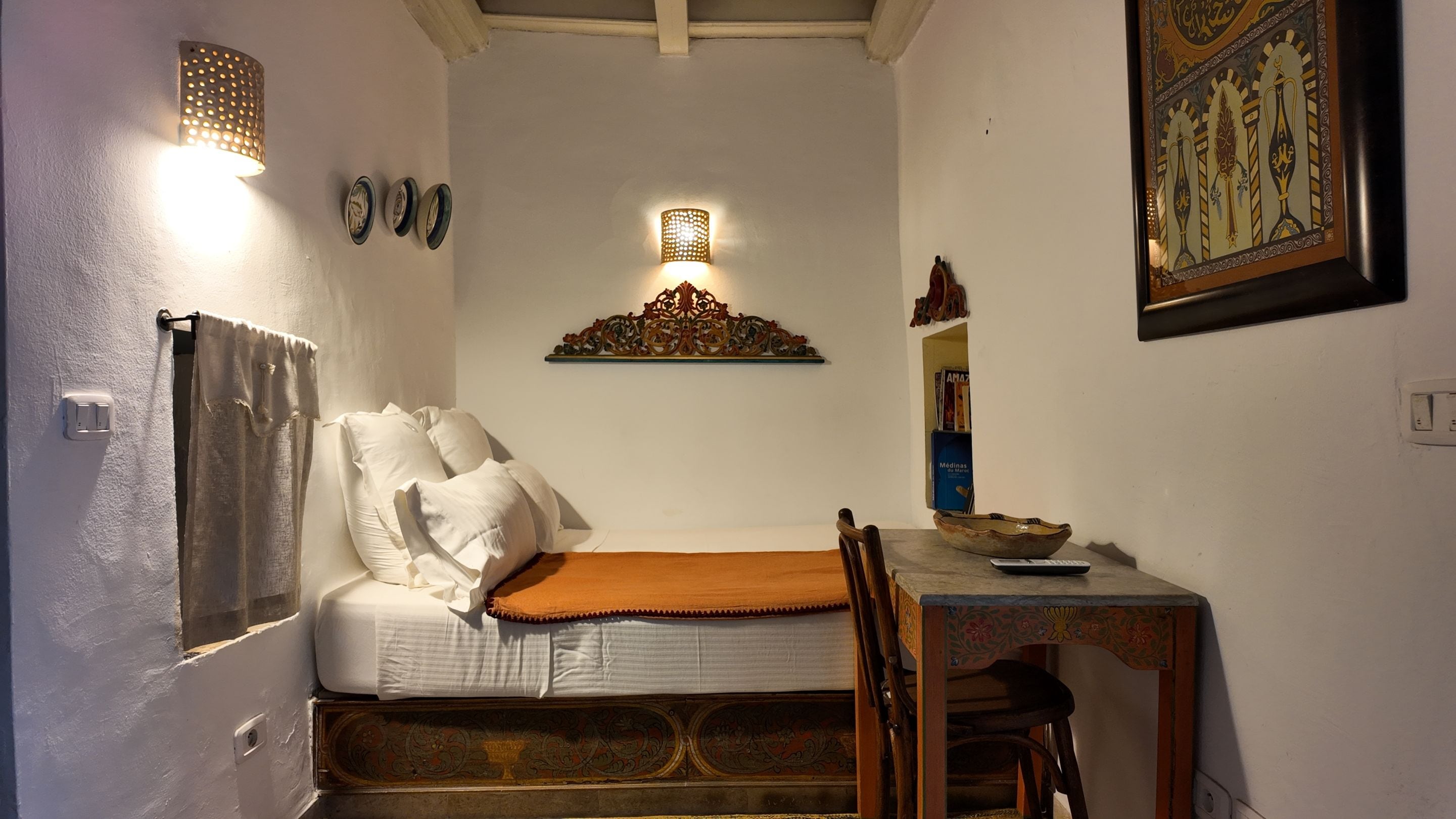 A small bedroom with a bed, a table with a bowl and a remote, a decorative wall piece, and a power outlet.