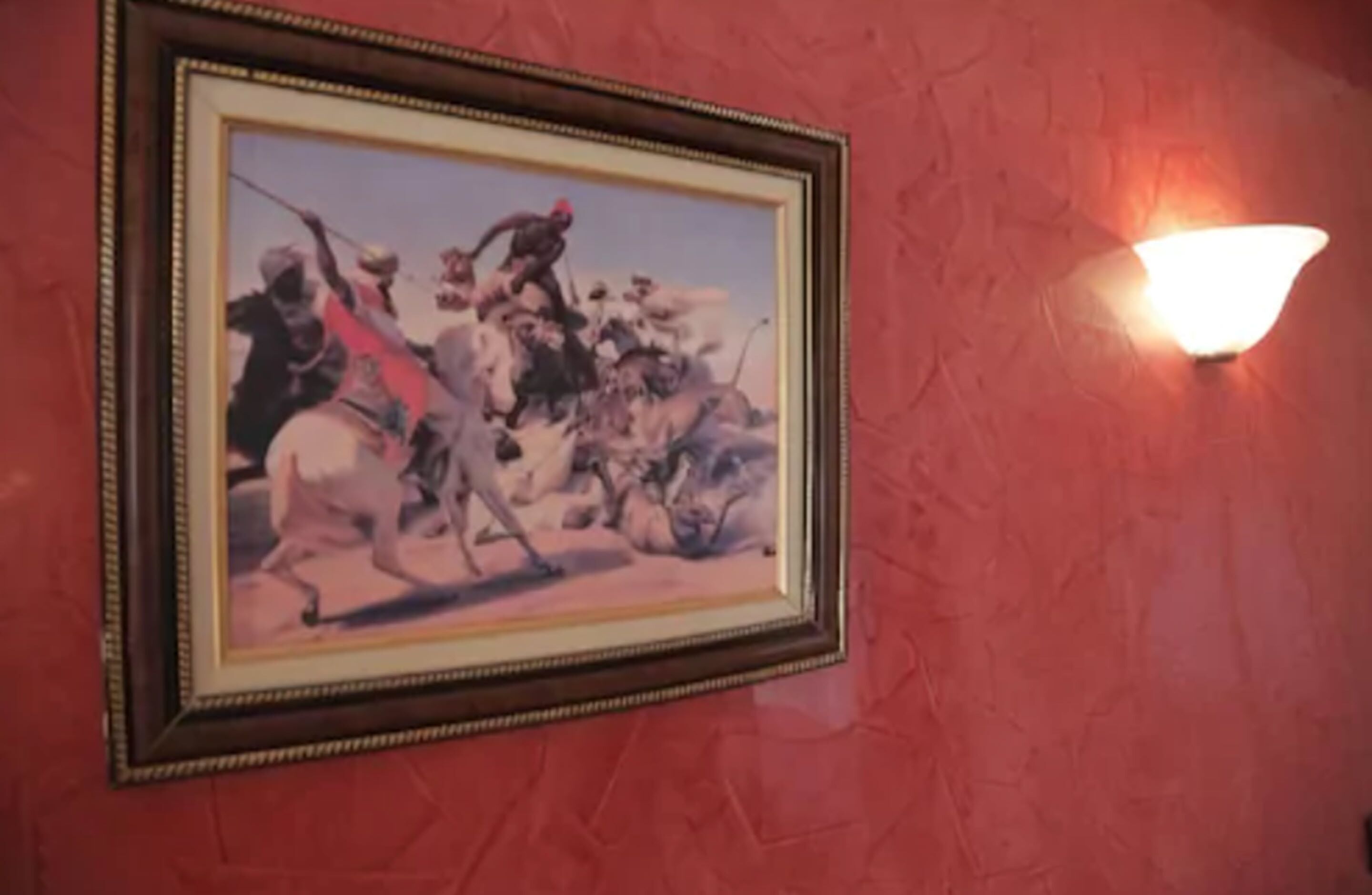 Escena de batalla histórica en un marco sobre una pared roja, con una lámpara de pared.