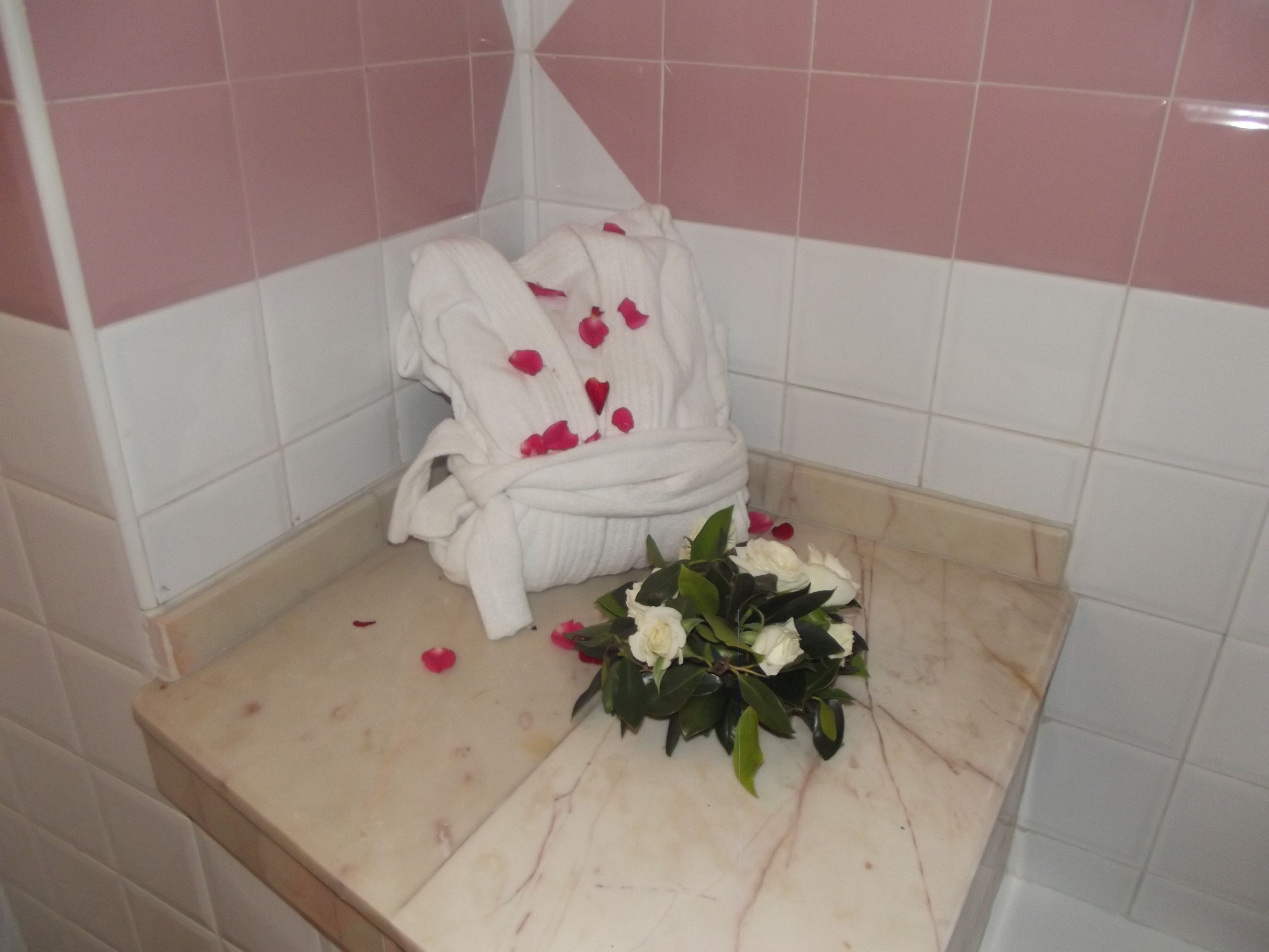 Un baño con una bañera blanca, paredes con azulejos rosa y blancos, y un albornoz blanco con pétalos de rosa sobre una encimera color beige.