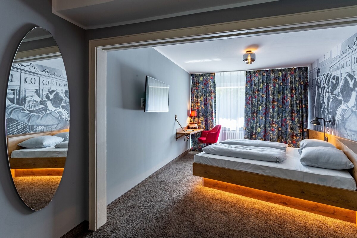 A hotel room with a bed, a desk, a chair, a mirror, and a window with curtains.