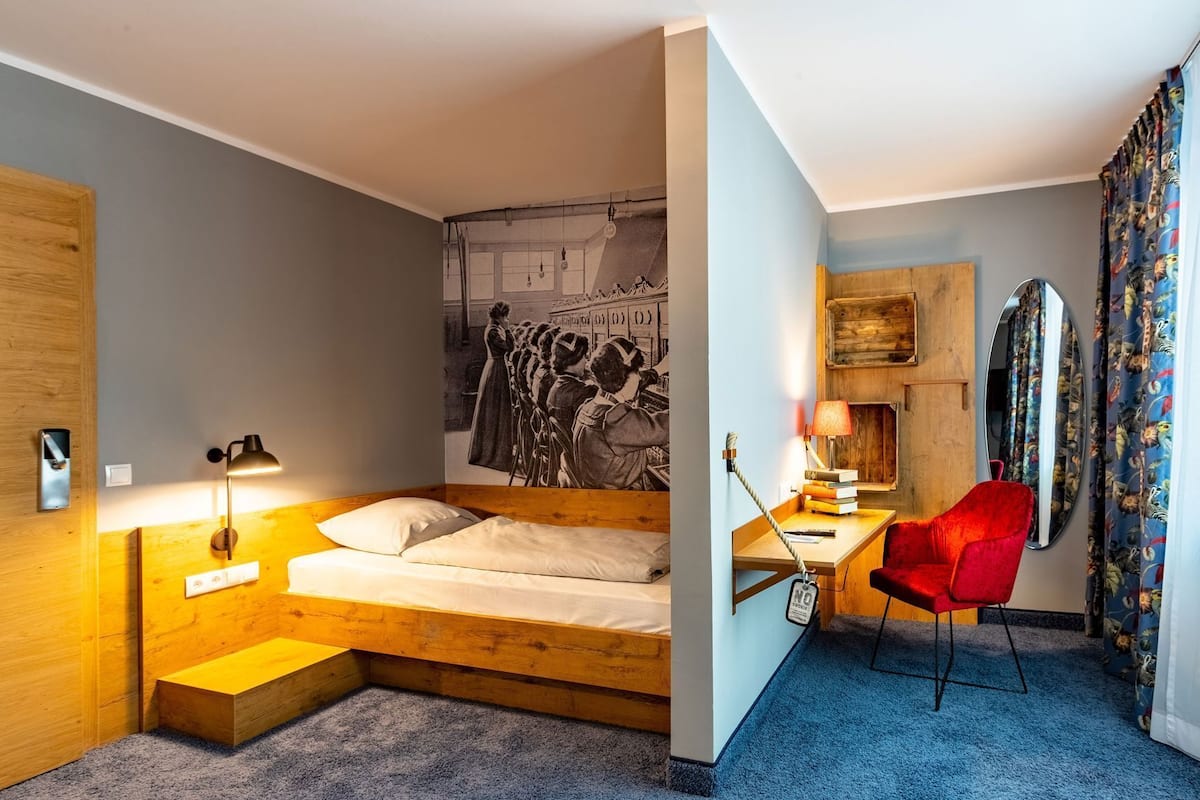 A room with a bed, a desk, a red chair, a wall with a black and white photo, and a blue carpet.