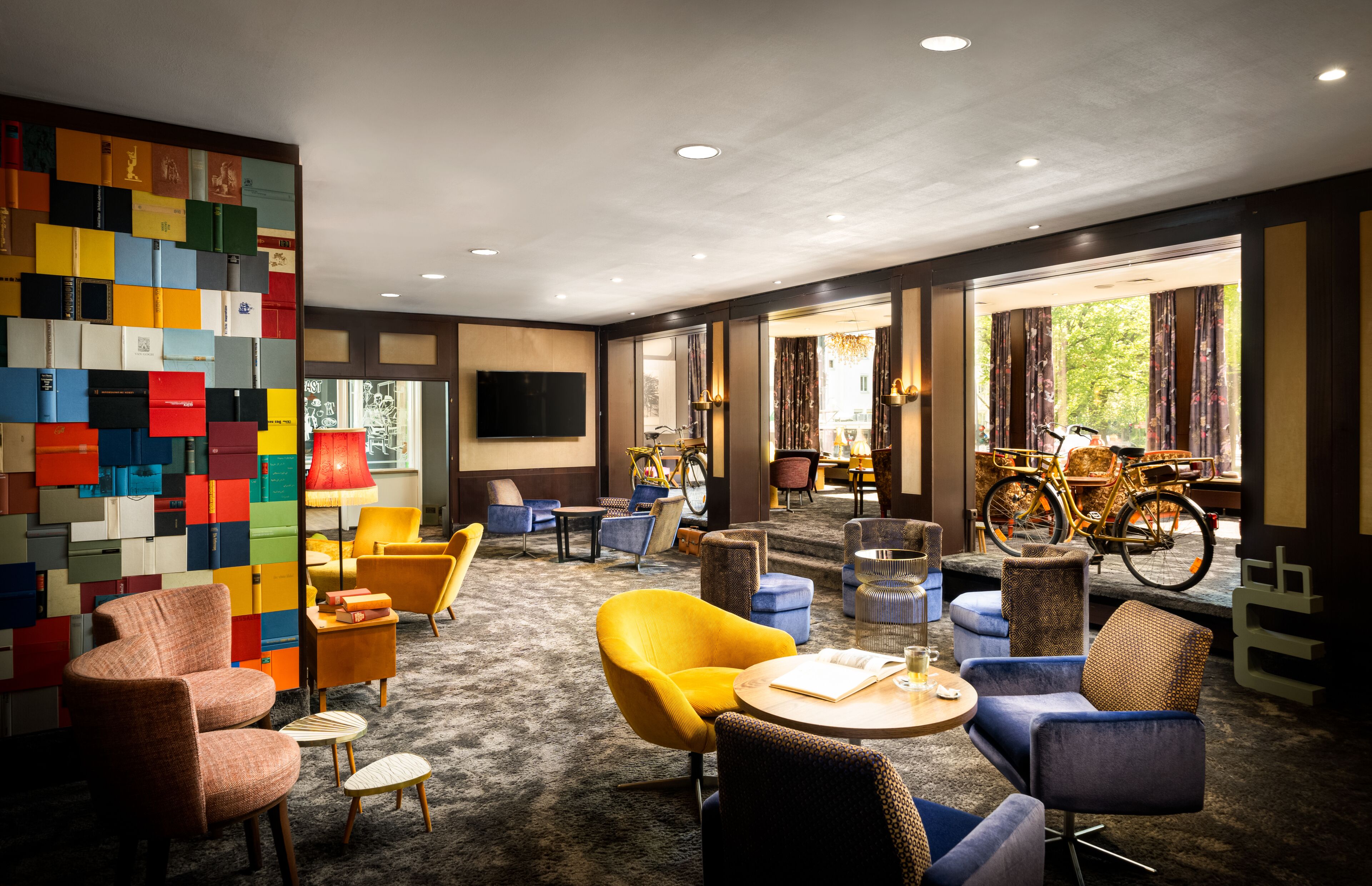 A modern hotel lobby with colorful block art on the wall, comfortable seating, a bicycle, and a large window with curtains.
