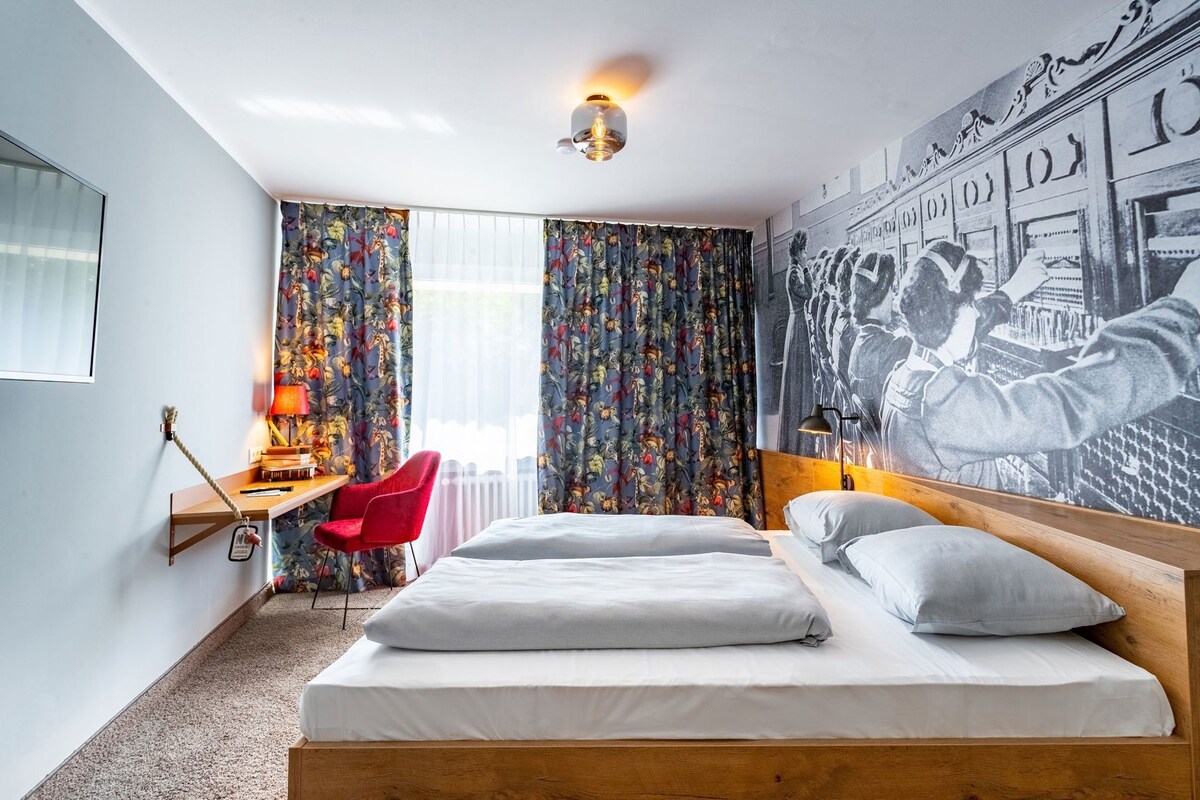 A hotel room with a bed, a desk, a chair, and a large wall mural.