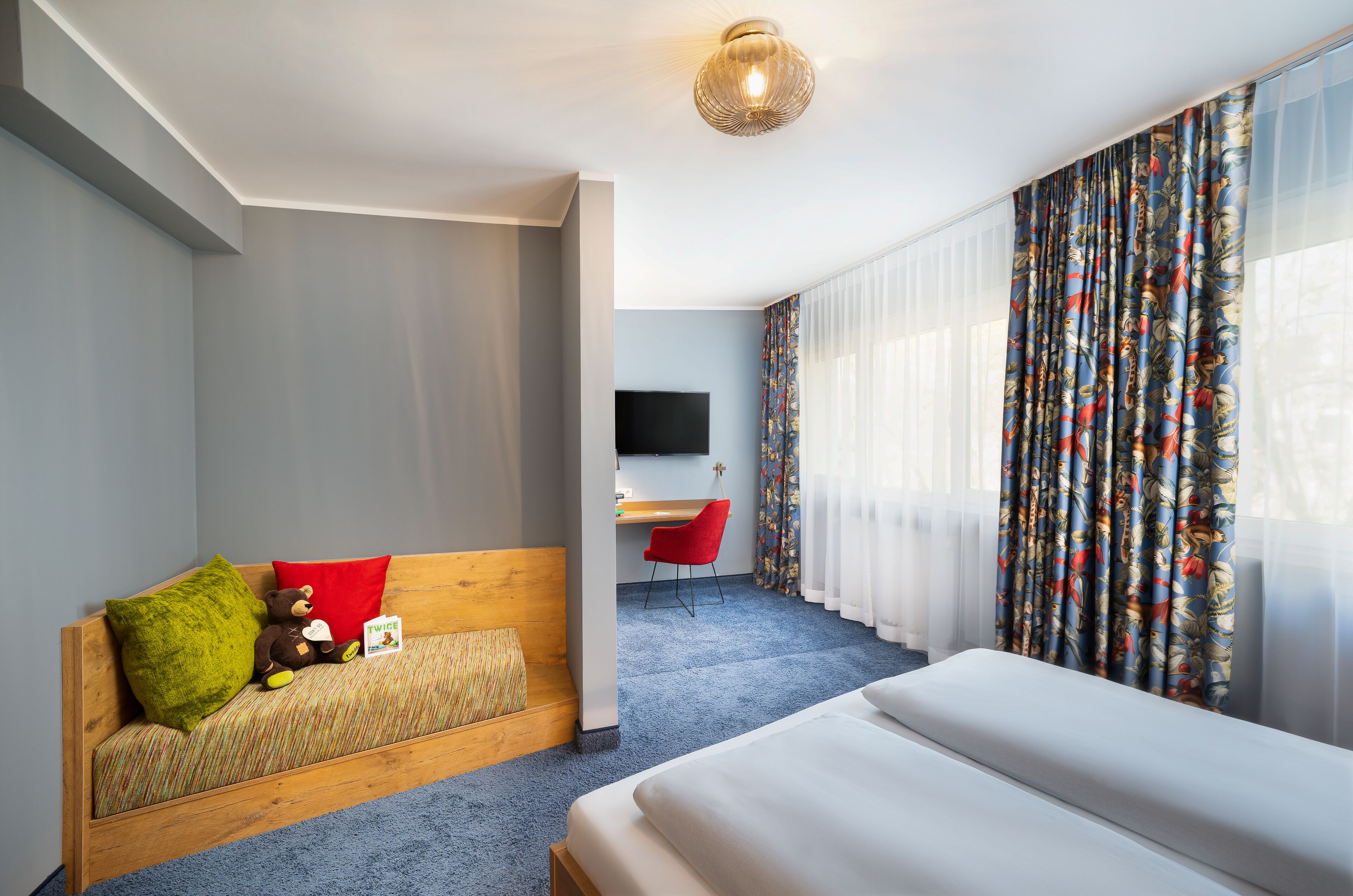 A modern hotel room with a wooden bed, a sofa bed with pillows, a red chair, a desk, and a TV.