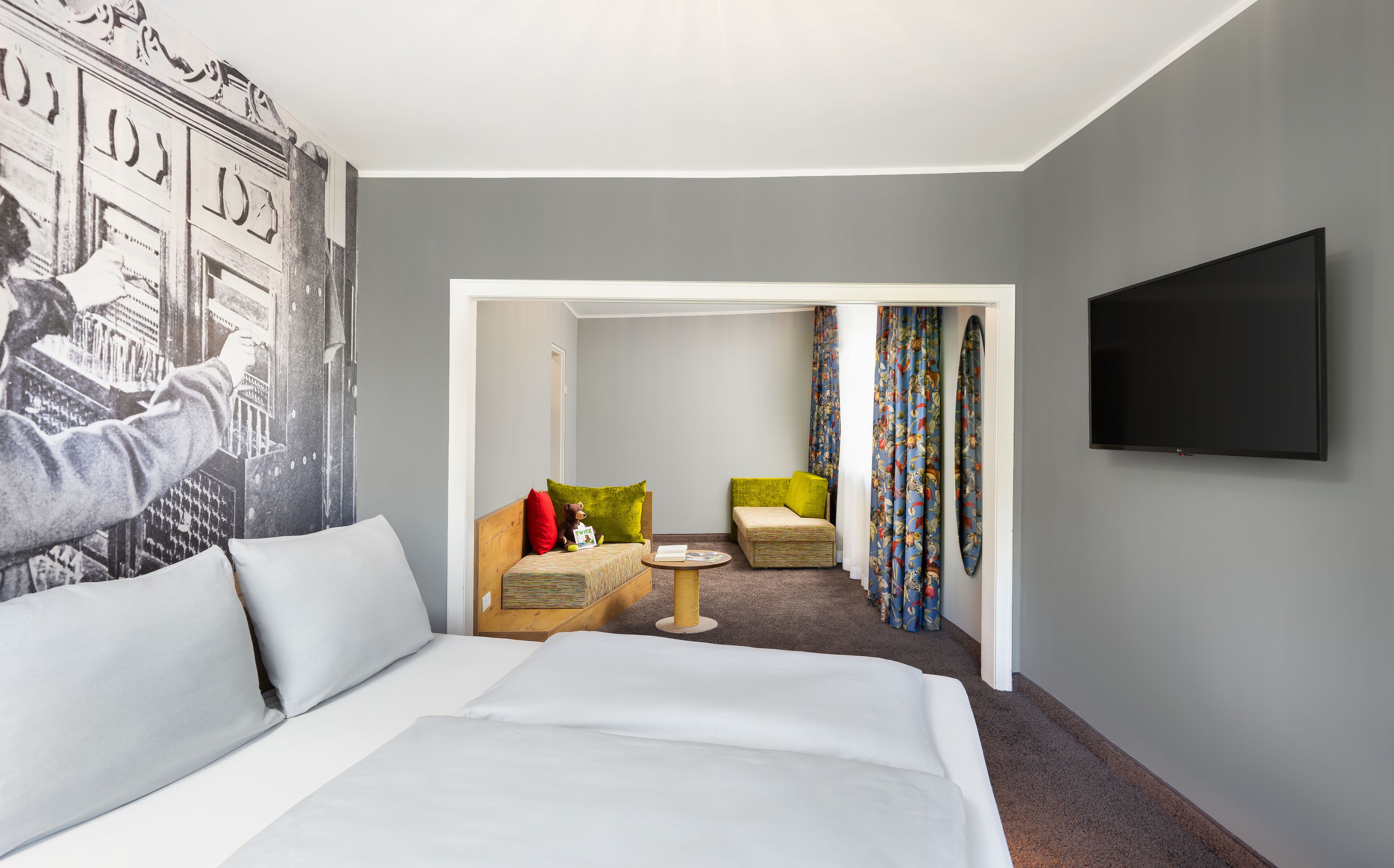 A hotel room with a bed, a TV, a seating area with chairs, and a colorful curtain.
