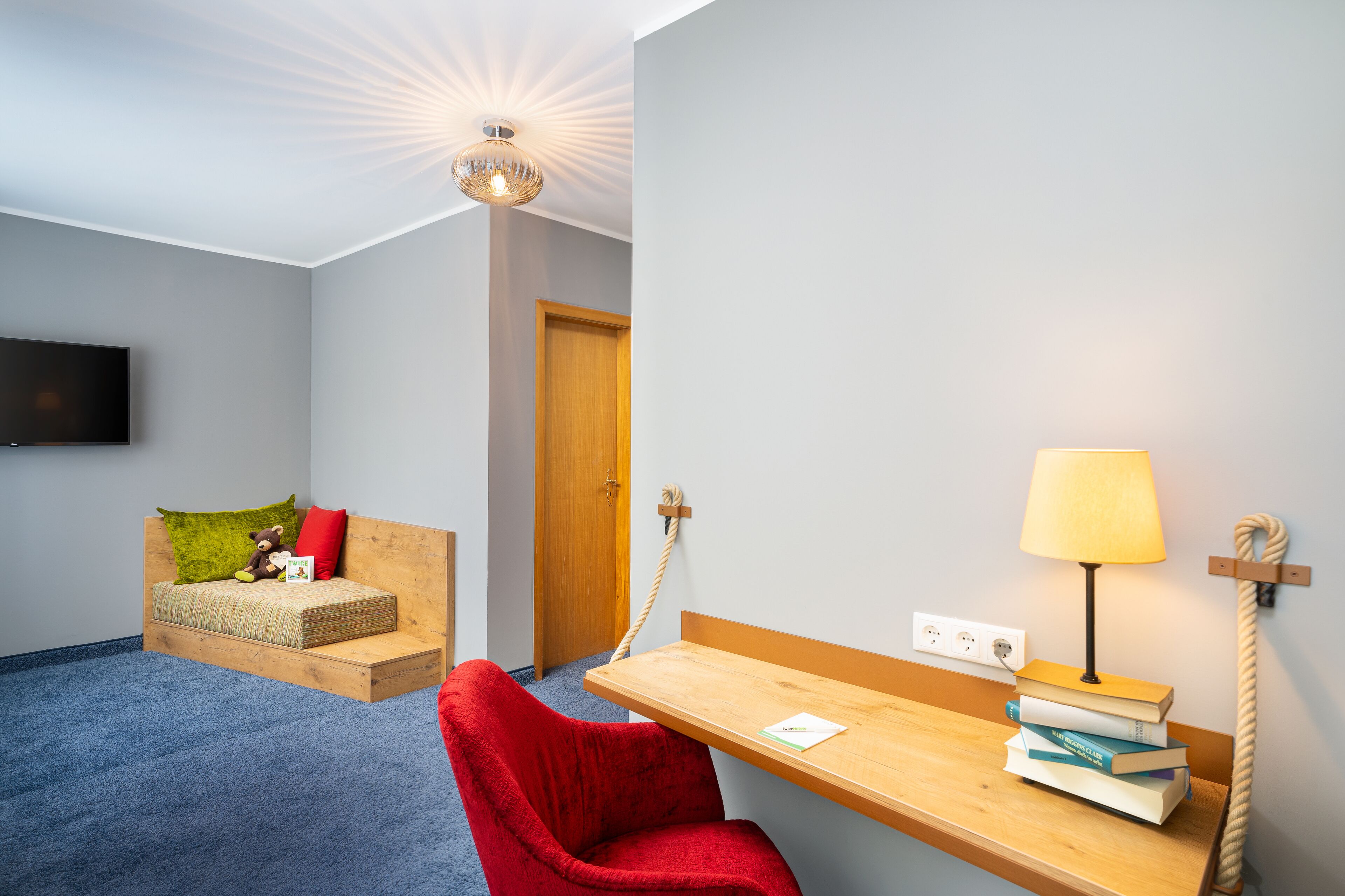 Une pièce comprenant un lit, un fauteuil rouge, un bureau en bois avec une lampe et un téléviseur fixé au mur.