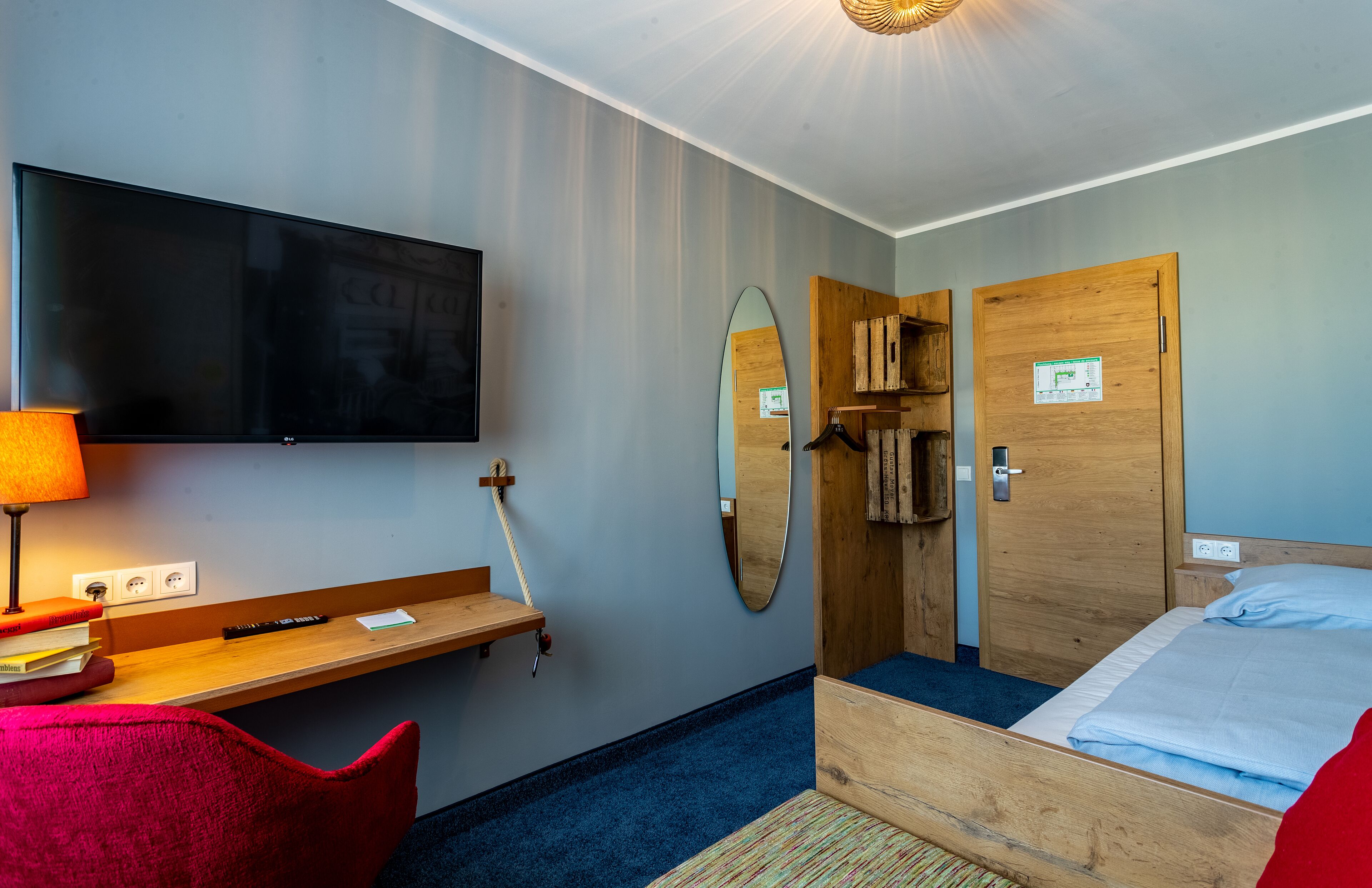 A hotel room with a bed, a desk with a lamp, a TV, a mirror, and a wooden door.
