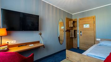 A hotel room with a bed, a desk with a lamp, a TV, a mirror, and a wooden door.