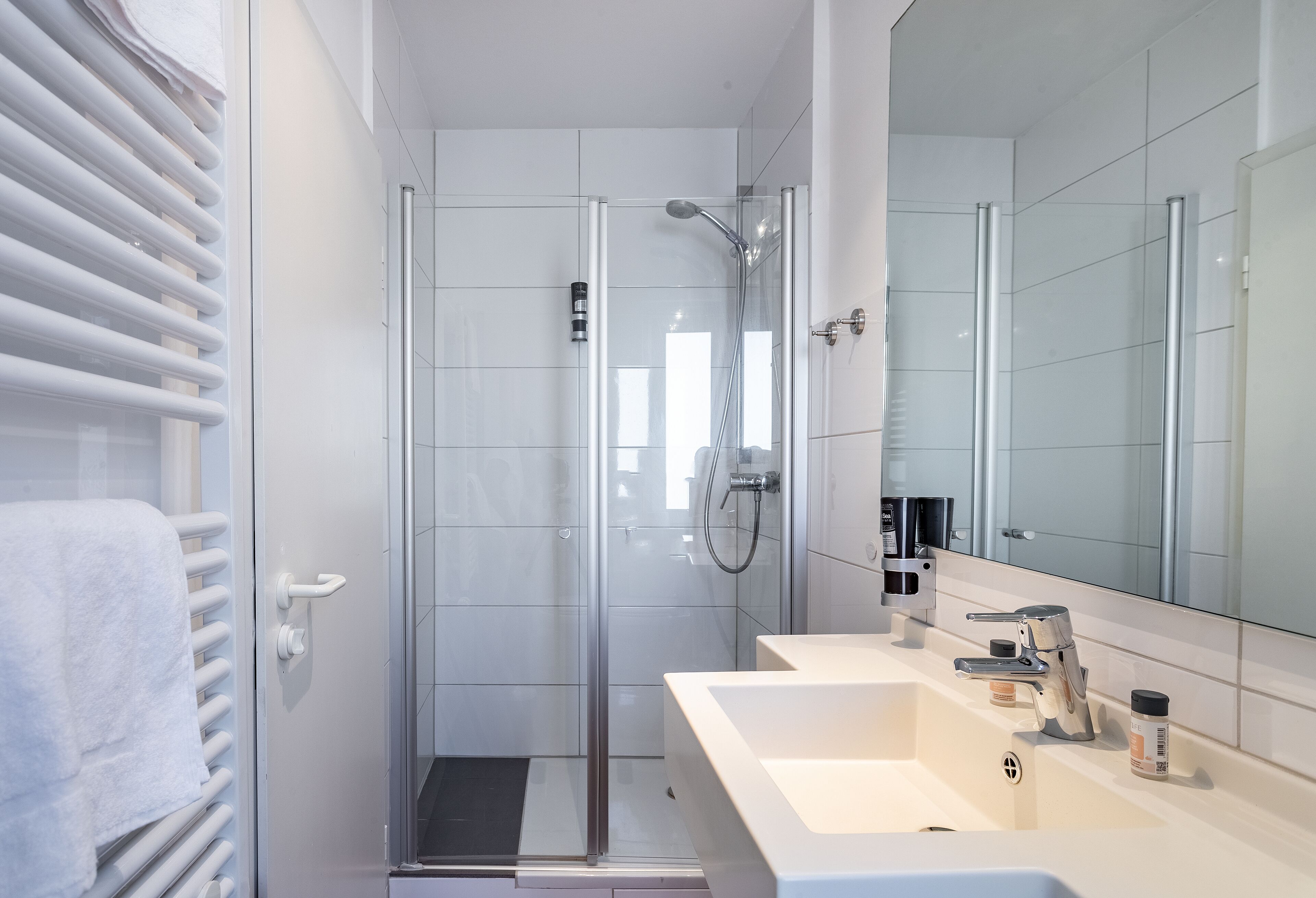 Un baño moderno con ducha envuelta en cristal, un lavabo blanco con espejo y un perchero para toallas.