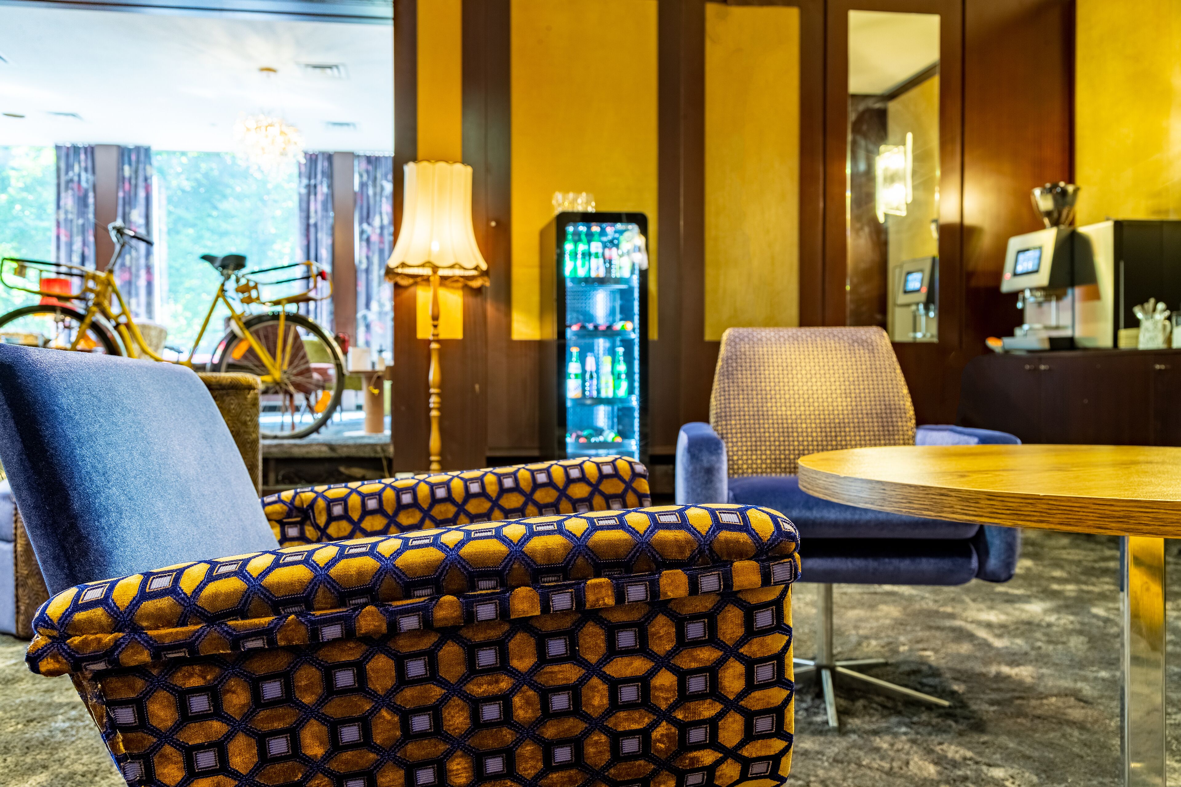 A room with patterned armchairs, a round table, a lamp, a bicycle, and a refrigerator.