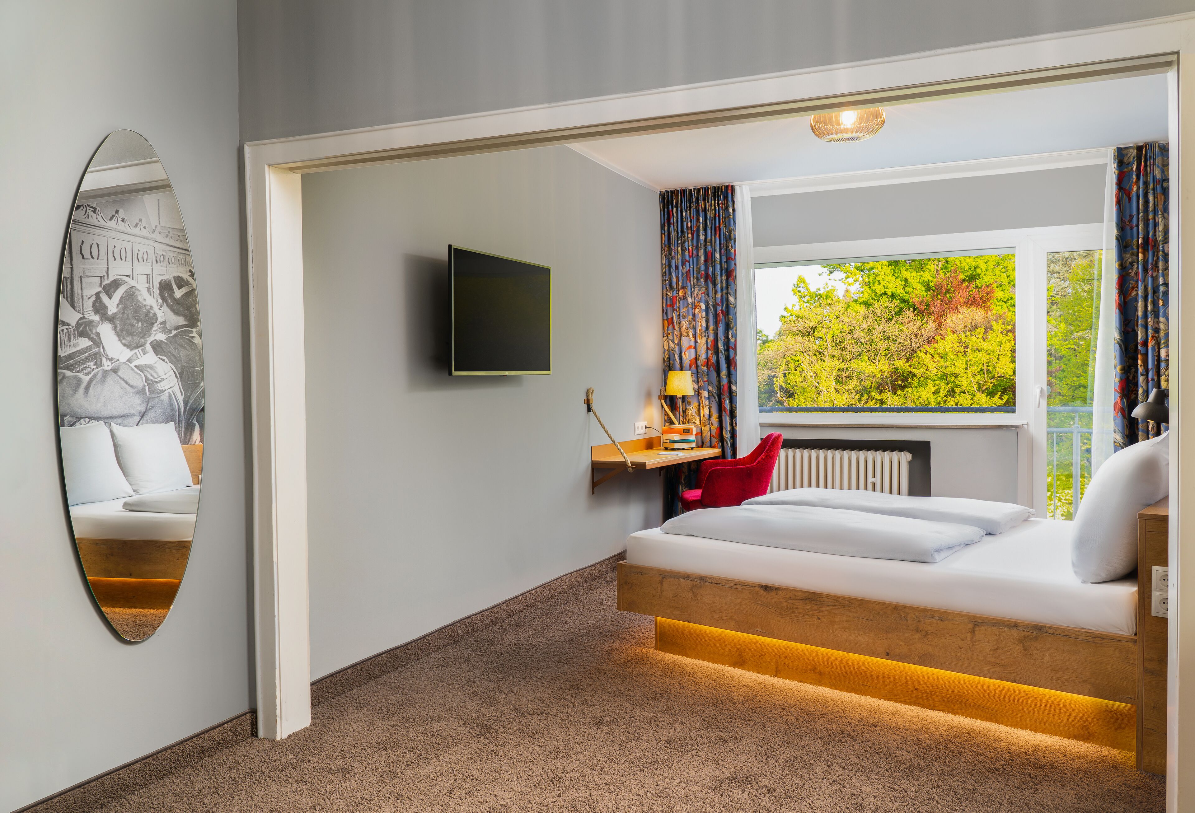 A modern hotel room with a bed, a TV, a desk with a lamp, and a view of trees through the window.