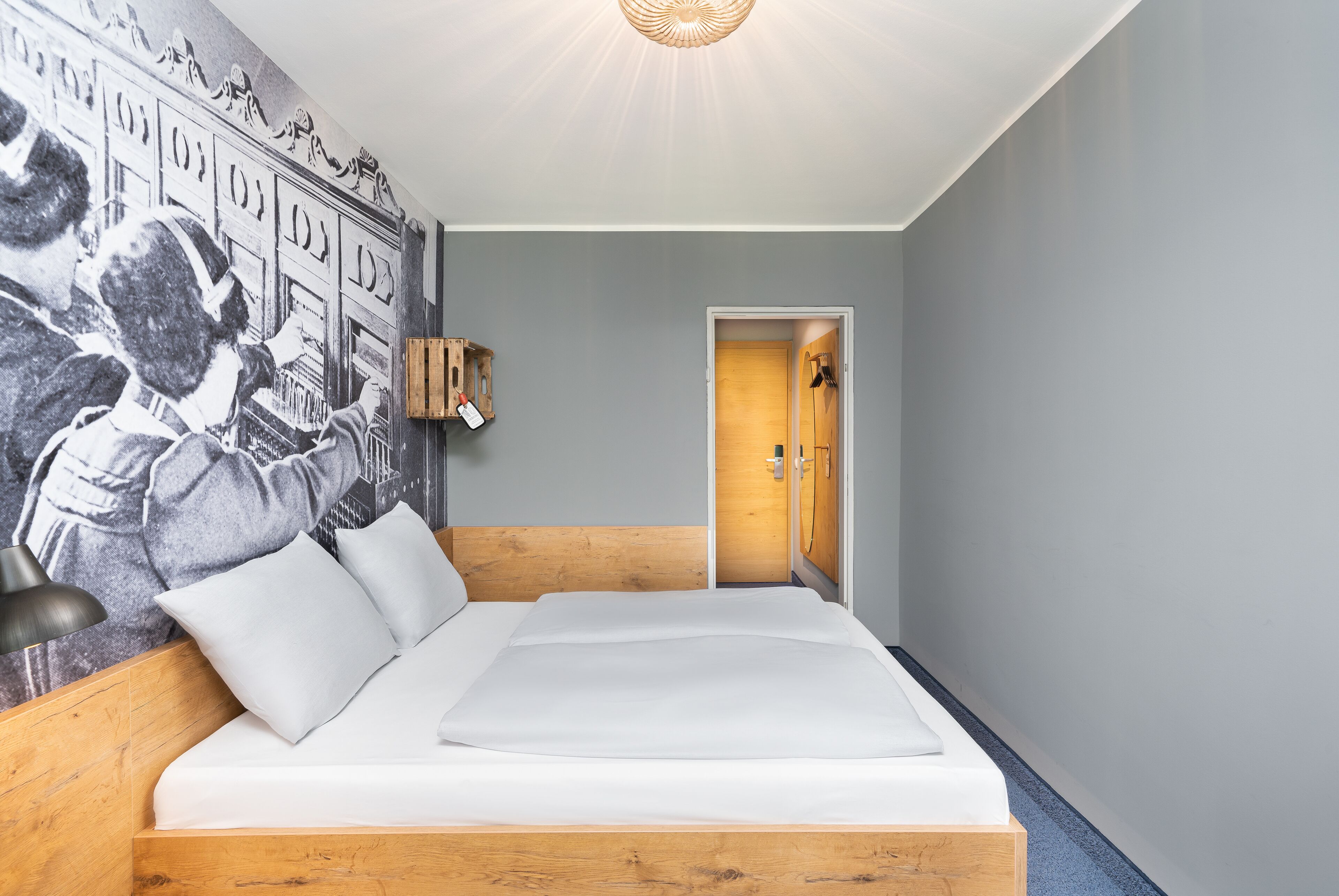 Une chambre à coucher avec une tête de lit en bois, un grand lit recouvert de linge de lit blanc, une lampe fixée au mur et une porte donnant sur une autre pièce.