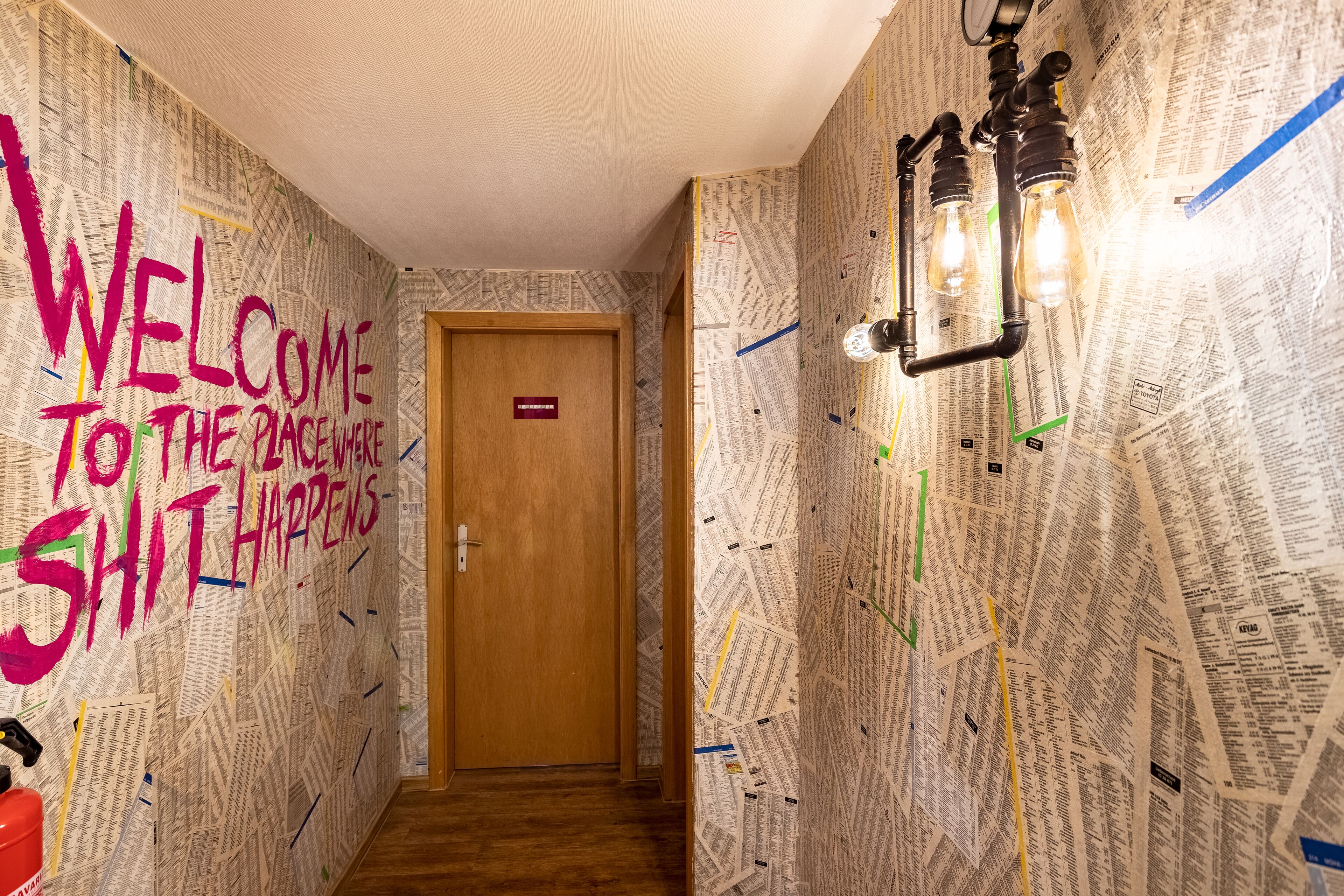 A hallway with a wooden door, walls covered in newspaper clippings, and a light fixture with exposed bulbs.
