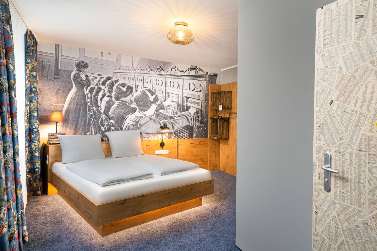 A bedroom with a wooden bed frame, a wall mural, a wooden wardrobe, and a door with a newspaper pattern.