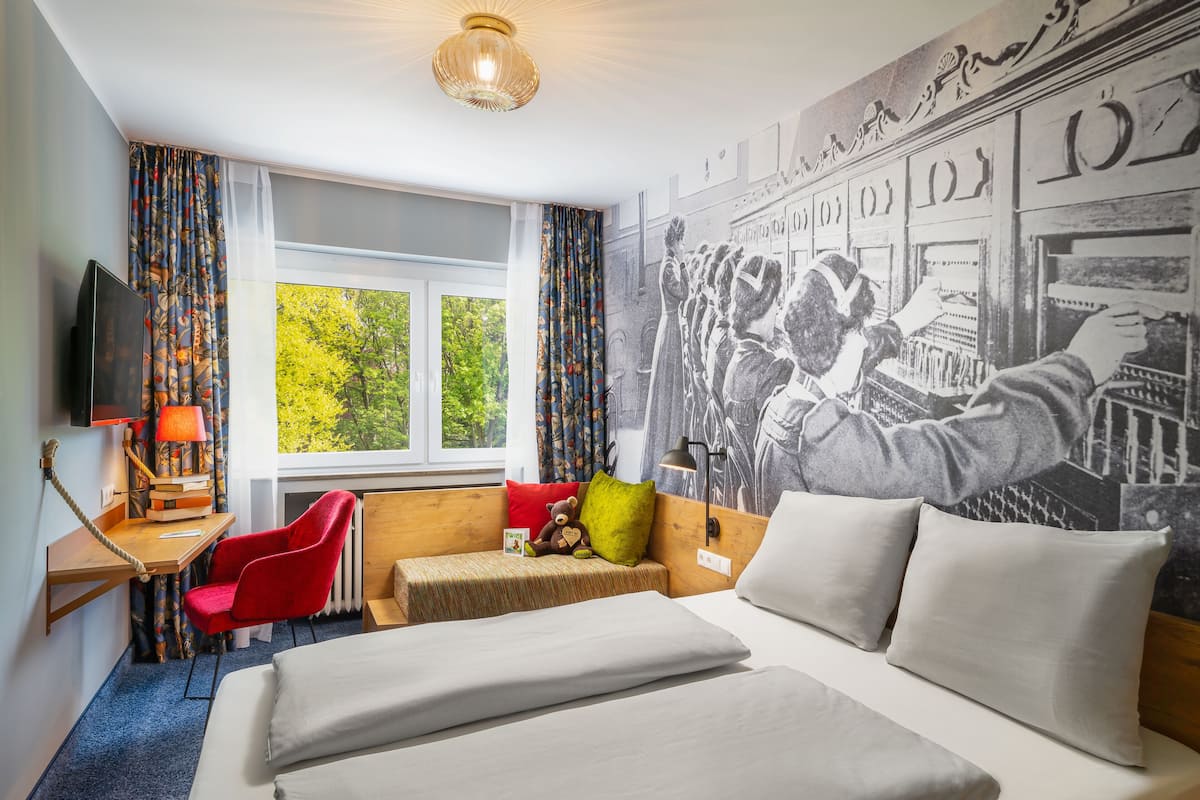 A hotel room with a bed, a desk with a lamp, a chair, a window with a view of trees, and a wall with a large black-and-white mural.