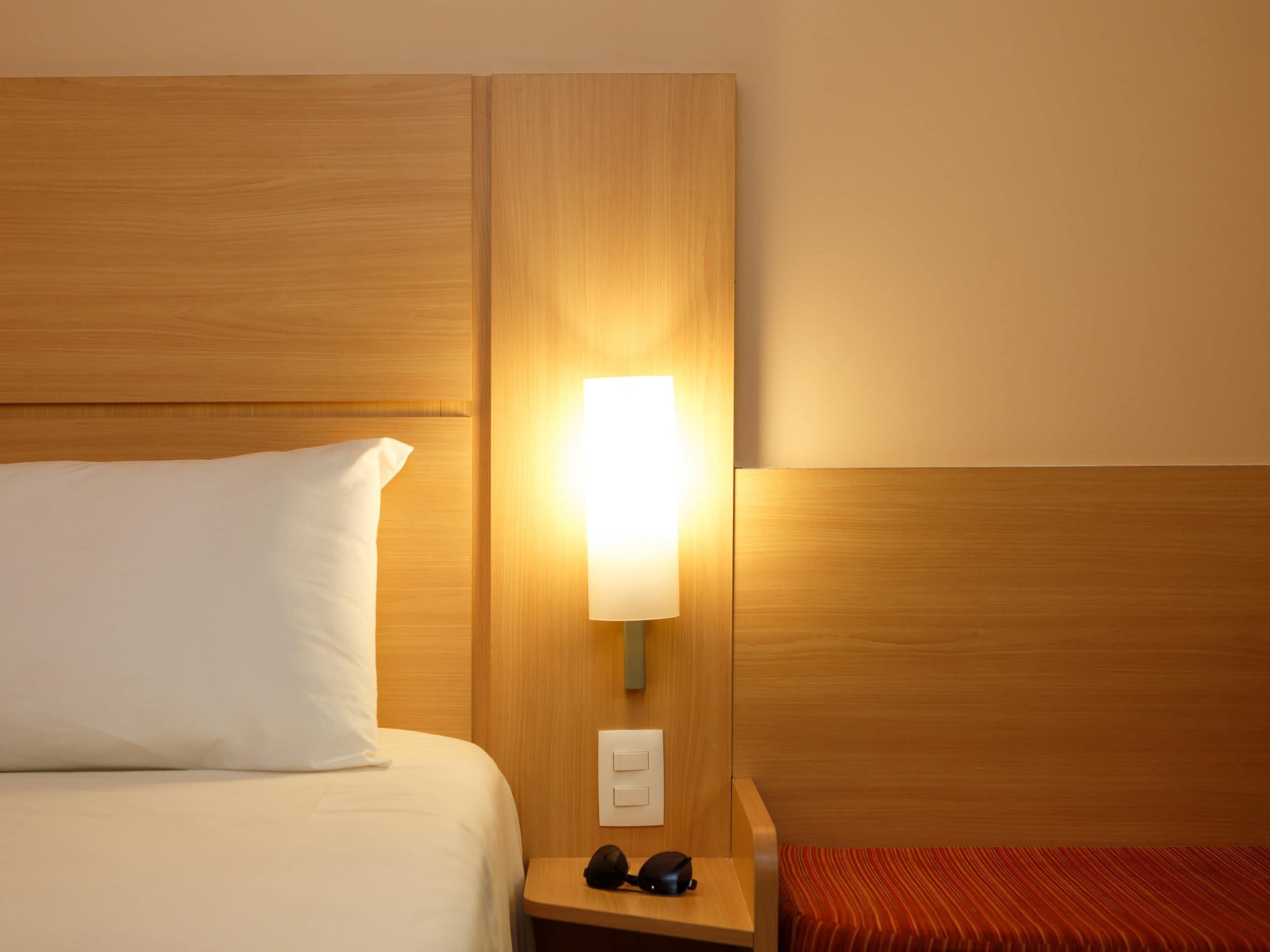 A hotel room with a bed, a bedside table with a lamp, and a power outlet.