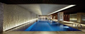 A long, narrow indoor swimming pool with a blue water surface, surrounded by stone walls and wooden paneling.