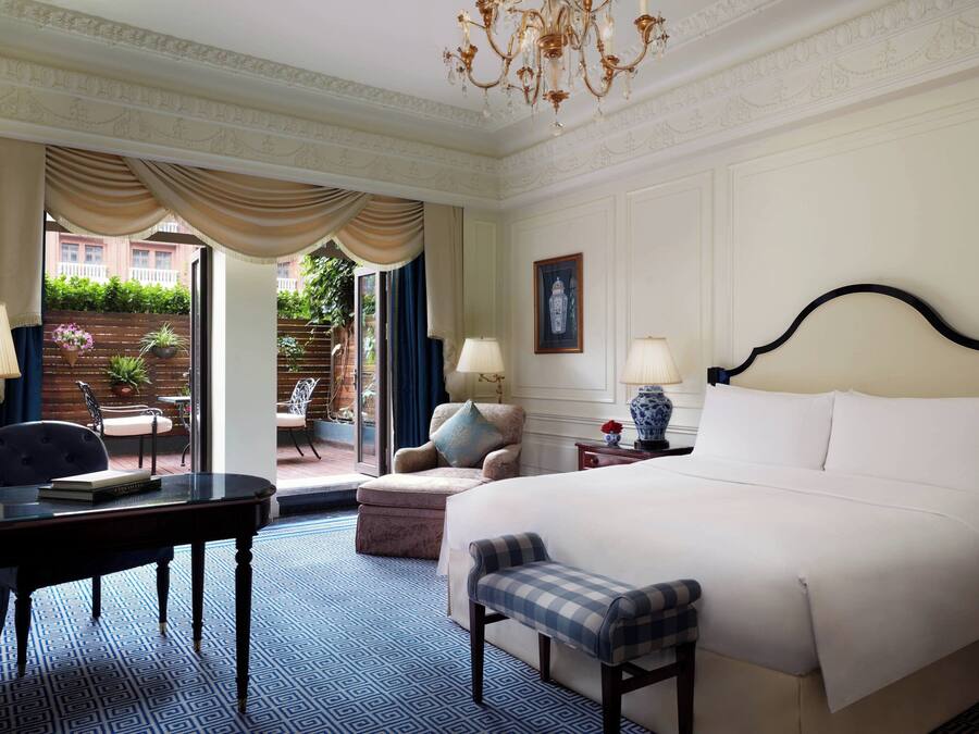 A hotel room with a large bed, a desk with a chair, a small table, and a bench. There is a chandelier, a window with curtains, and a door leading to an outdoor area with plants.