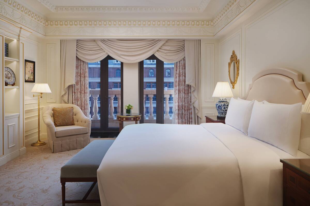 A hotel room with a large bed, a chair, a lamp, and a view of a building through a window.