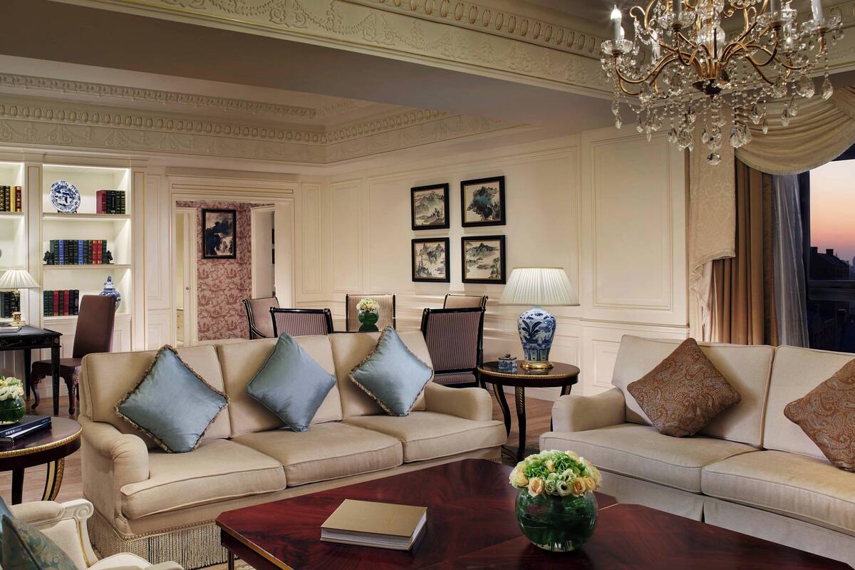 A luxurious living room with two sofas, a coffee table, and a chandelier. There are bookshelves and framed pictures on the walls.