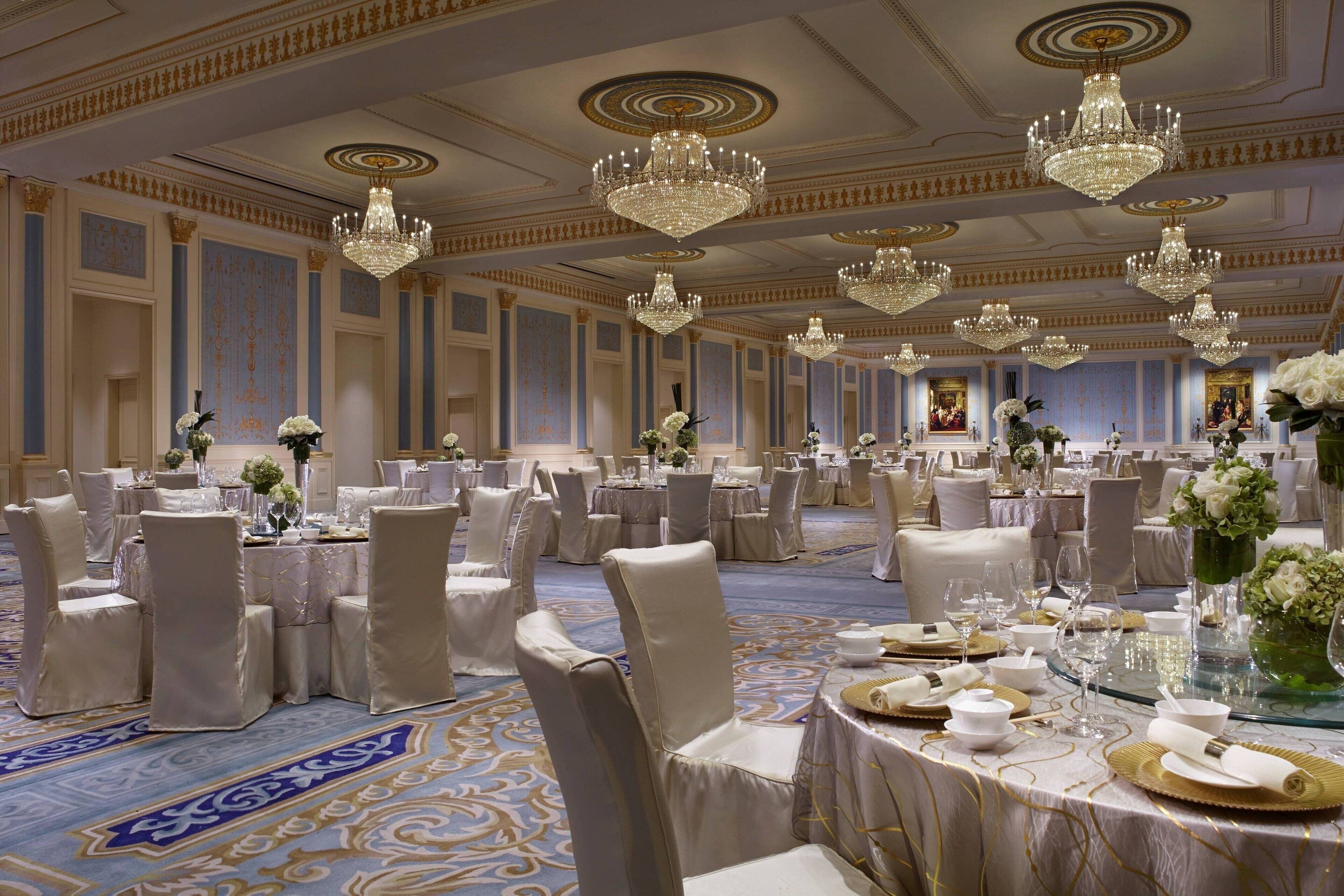 A banquet hall with round tables, white chairs, and chandeliers.