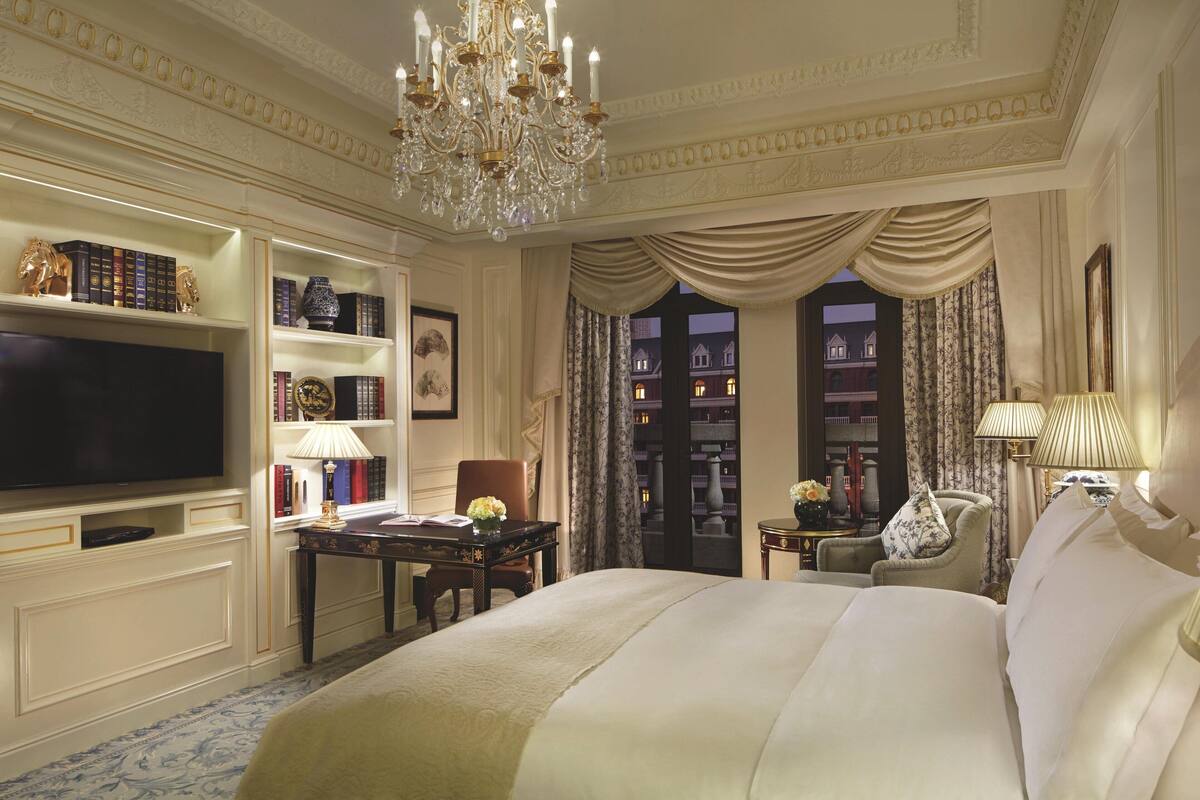 A luxurious hotel room with a large bed, a television, a bookshelf, and a chandelier.
