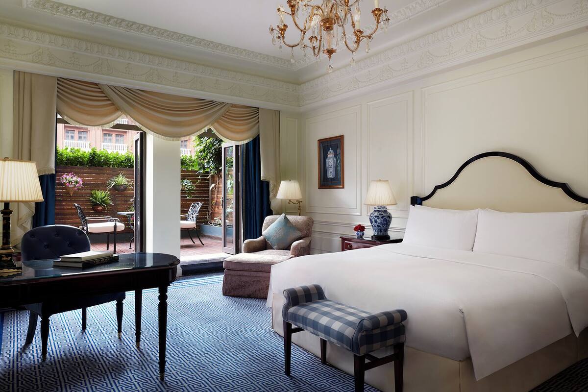 A hotel room with a large bed, a desk with a chair, a bench, a chandelier, and a view of the outside.