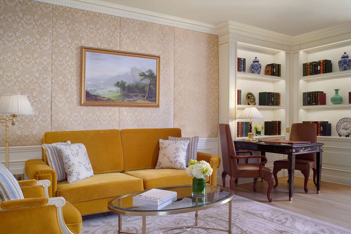 A room with a yellow sofa, a glass coffee table, a painting, and a bookshelf with books and decorative items.