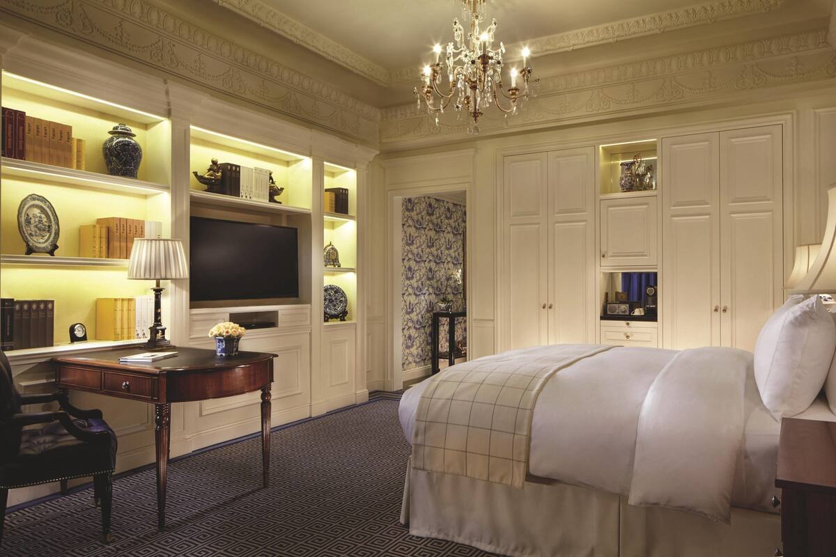 A hotel room with a large bed, a desk with a lamp, a television, and shelves with decorative items.